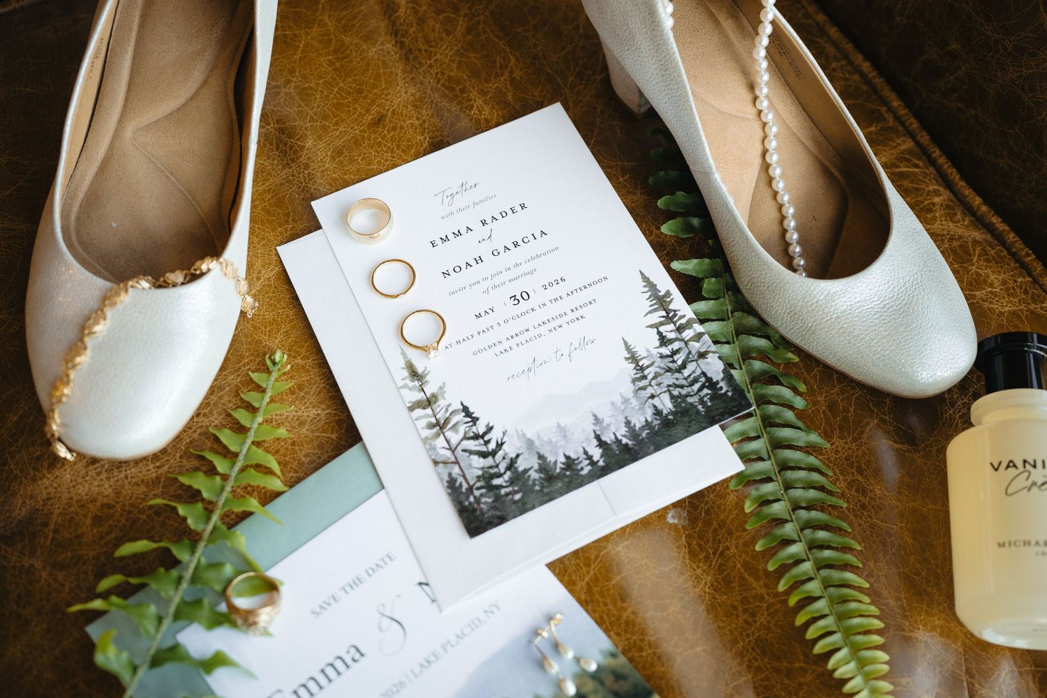 Wedding flat lay with forest-themed invitation, gold rings, white heels, and greenery.