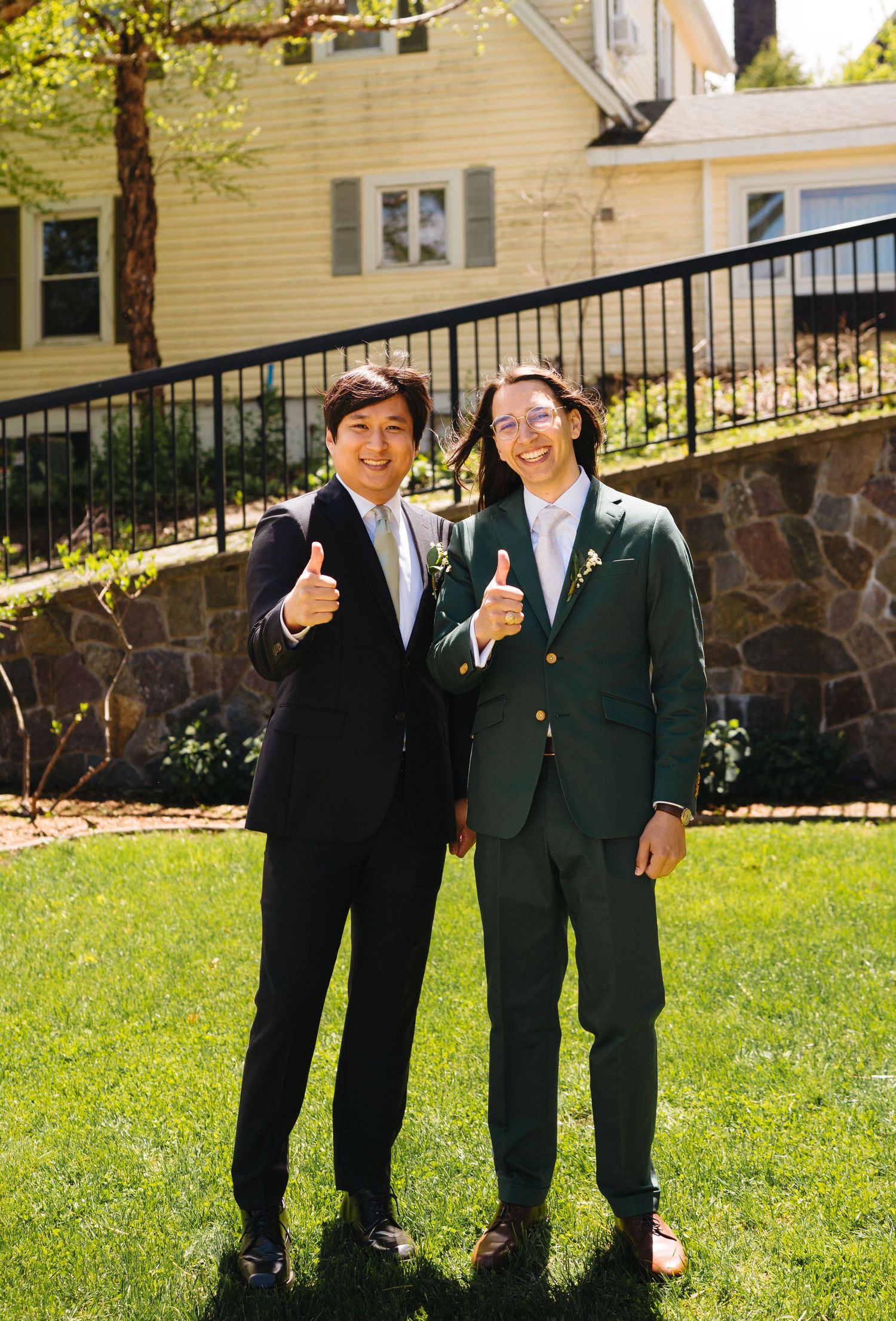 Two men in dark suits giving thumbs up while posing on a sunny lawn in front of a yellow house.