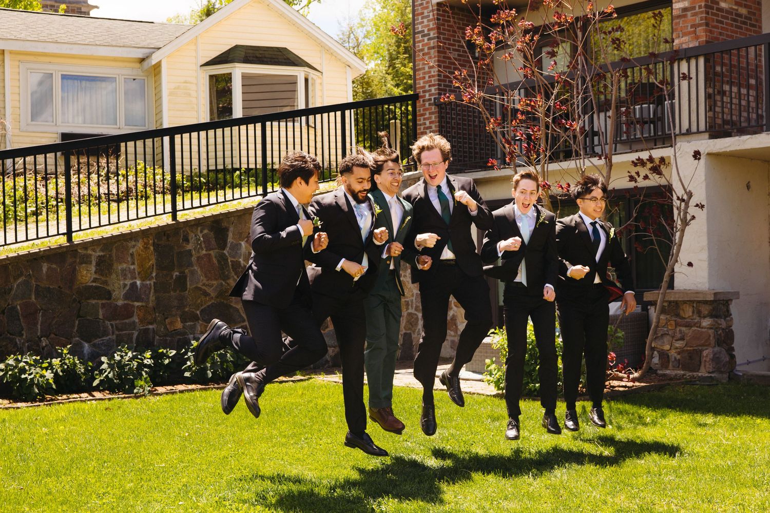 Excited groomsmen in black suits jumping joyfully on green lawn outside wedding venue.