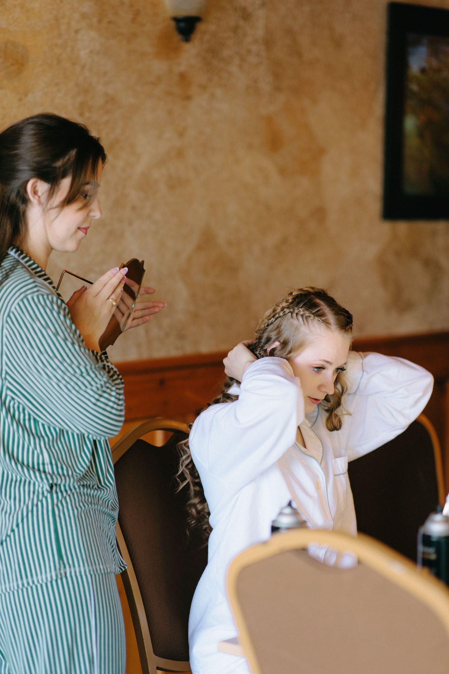 Bridesmaid helping bride with braided updo hairstyle during wedding morning preparations.