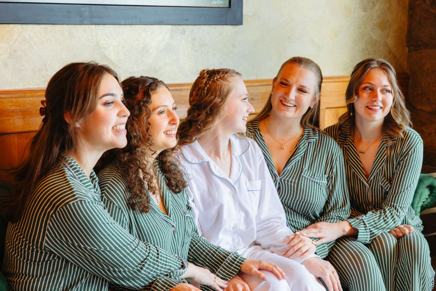 Bride in white robe laughing with bridesmaids in green striped pajamas during bridal getting ready session.