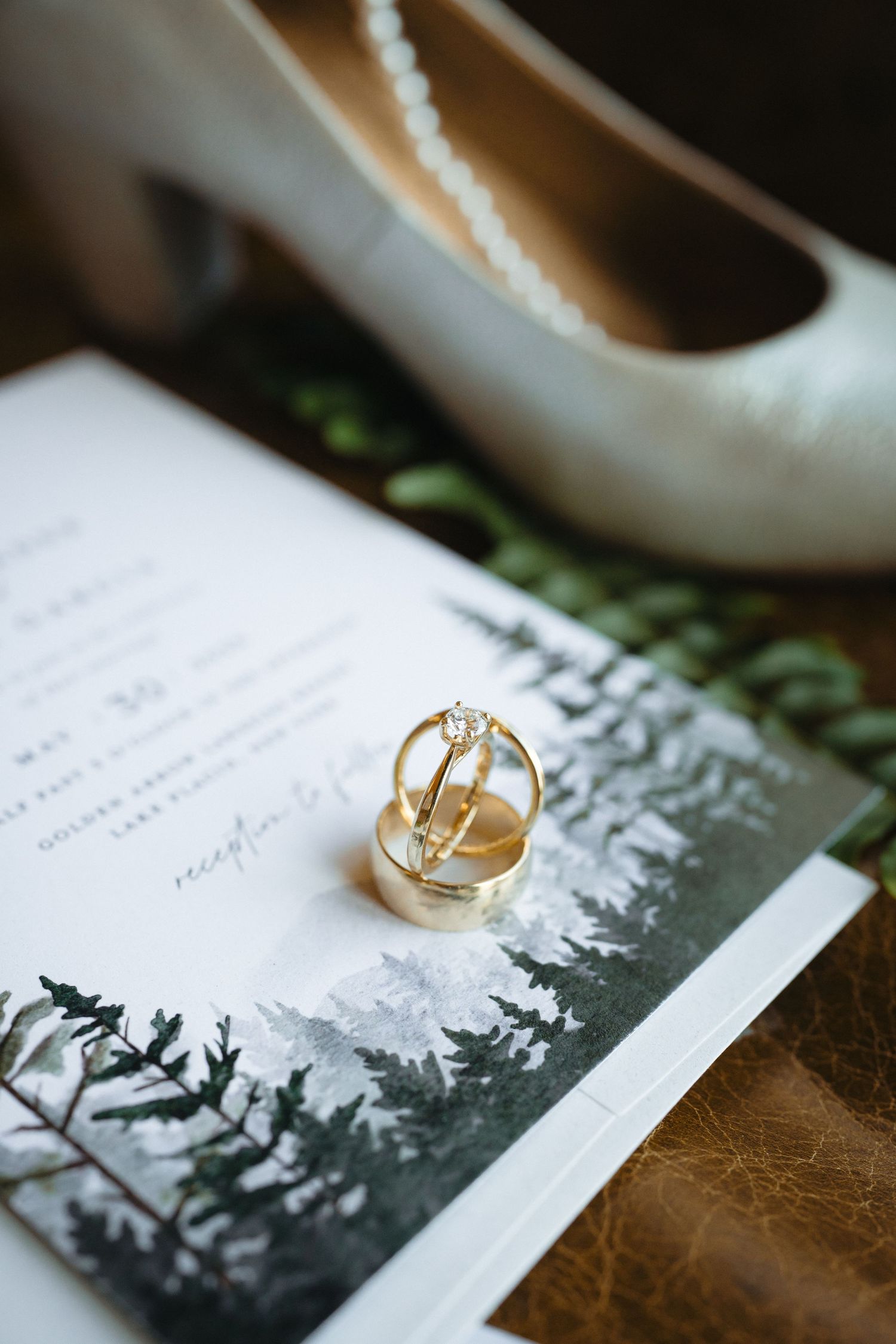 Gold wedding rings and diamond engagement ring displayed on pine tree watercolor wedding invitation with white heels.