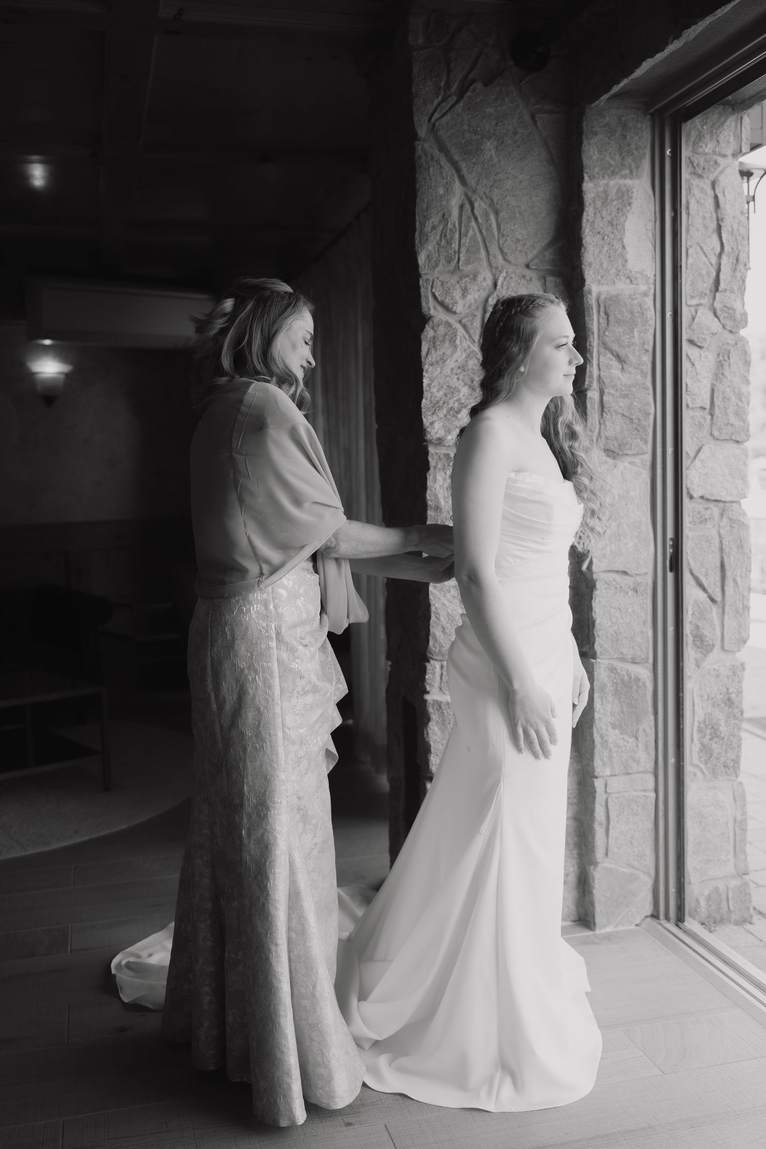 Black and white photo of bridesmaid helping bride with wedding dress near stone wall and window.