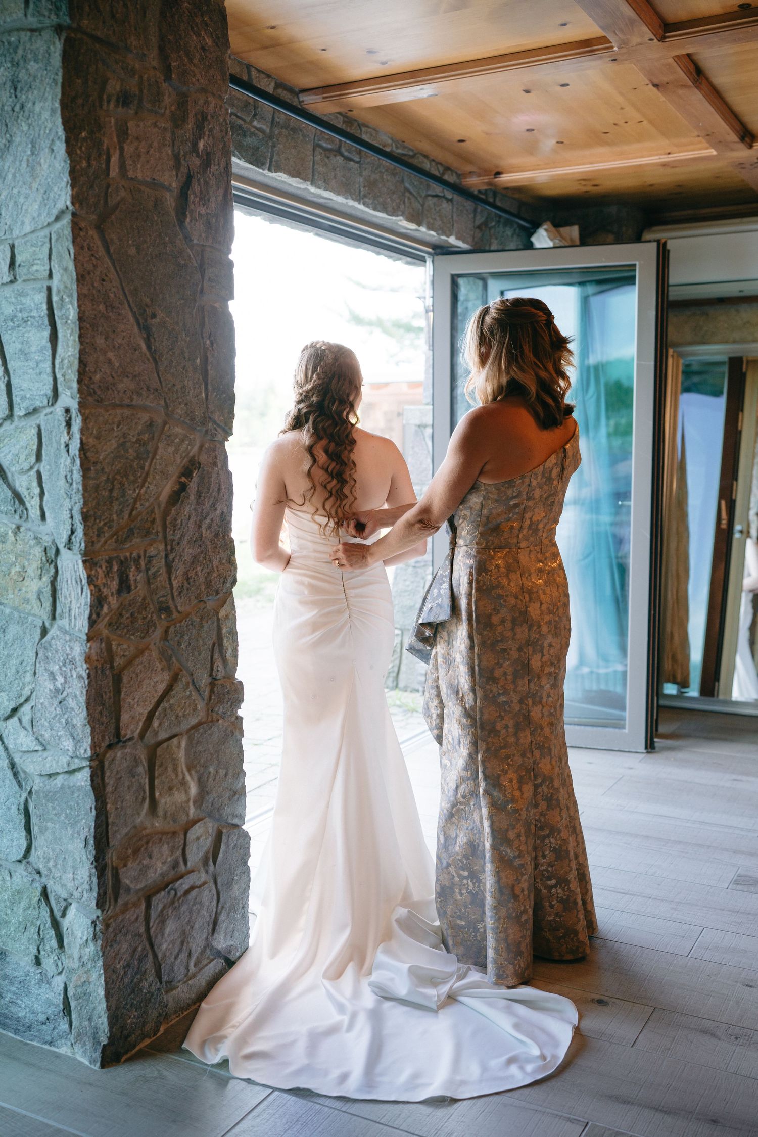 Bridesmaid helping bride with wedding dress zipper in rustic stone venue doorway.