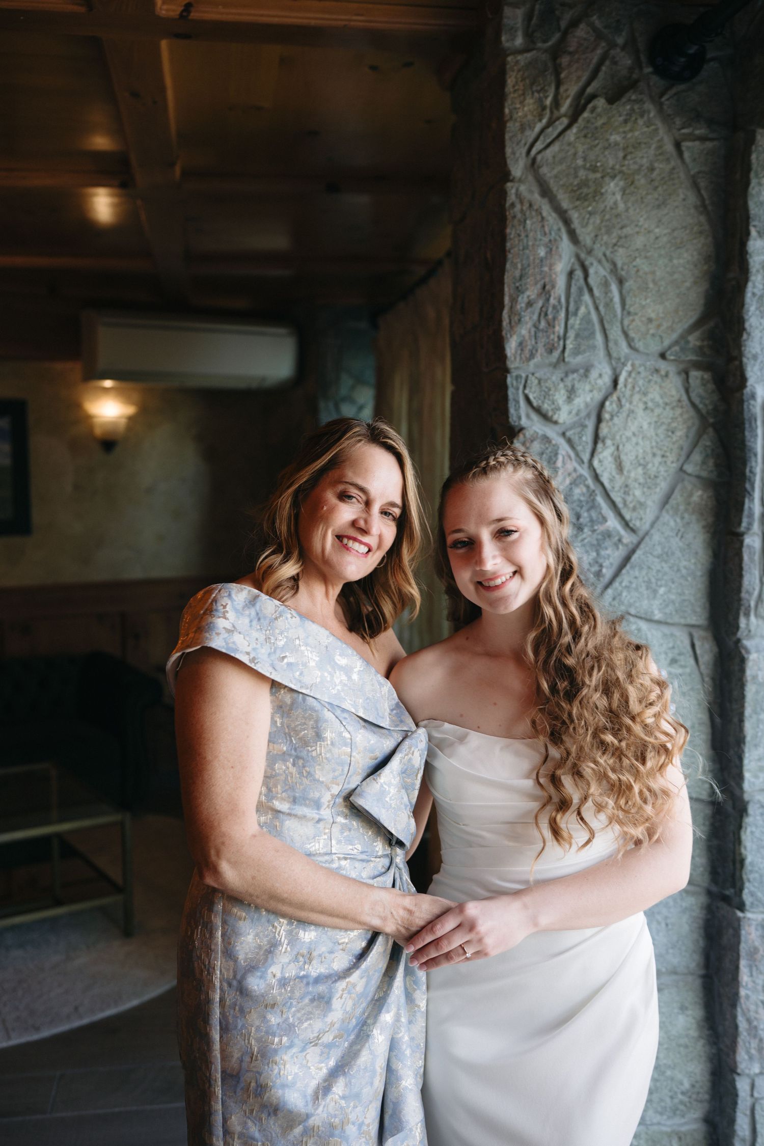Smiling bride in white strapless gown posing with older woman in blue floral dress against stone wall.