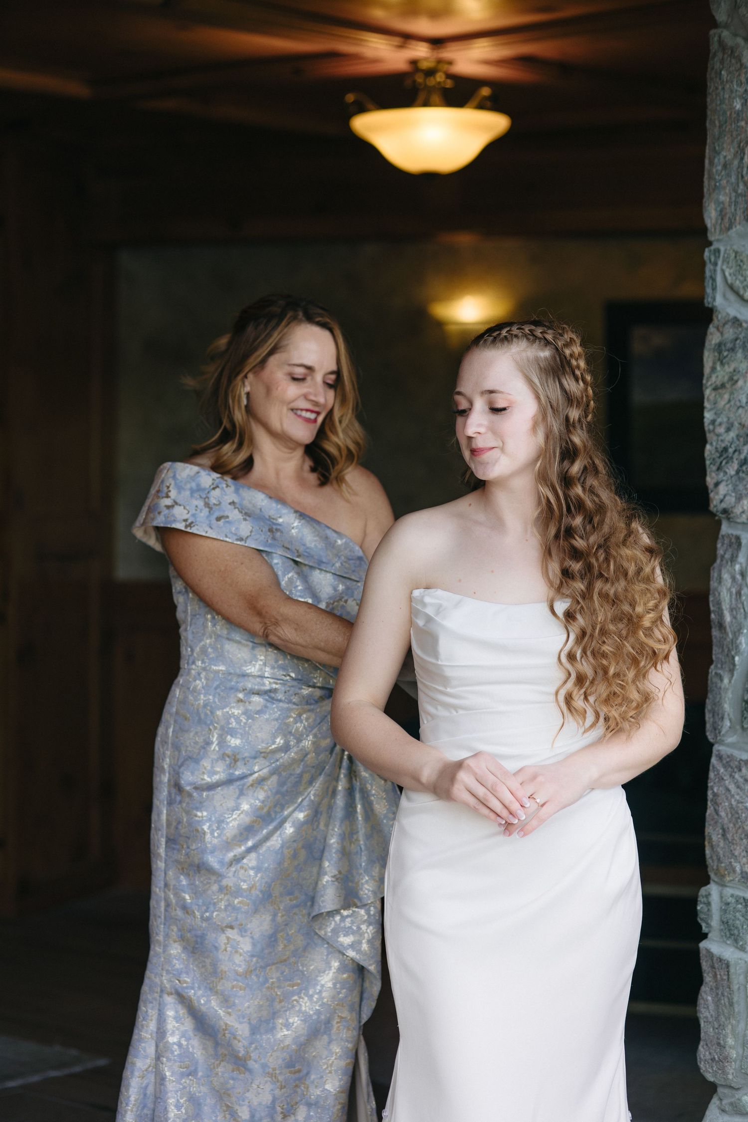 Mother in silver gown helping bride in strapless white wedding dress get ready in a rustic venue.