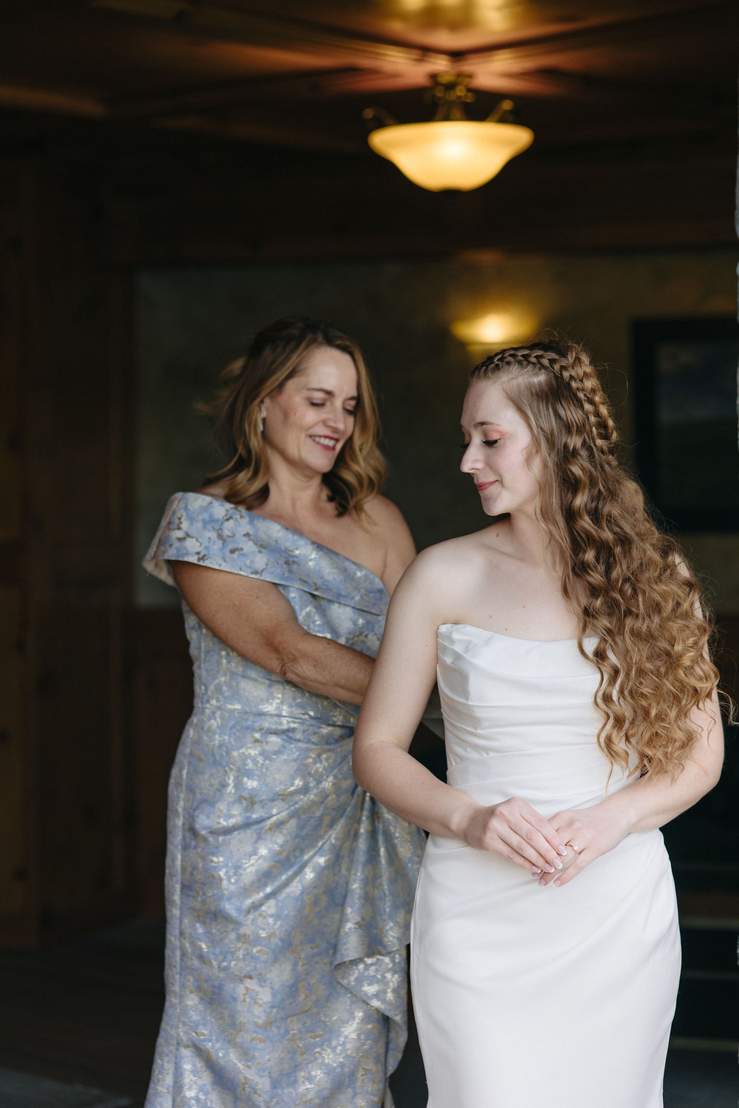 Mother in blue floral gown smiling at bride in strapless white wedding dress inside rustic venue.