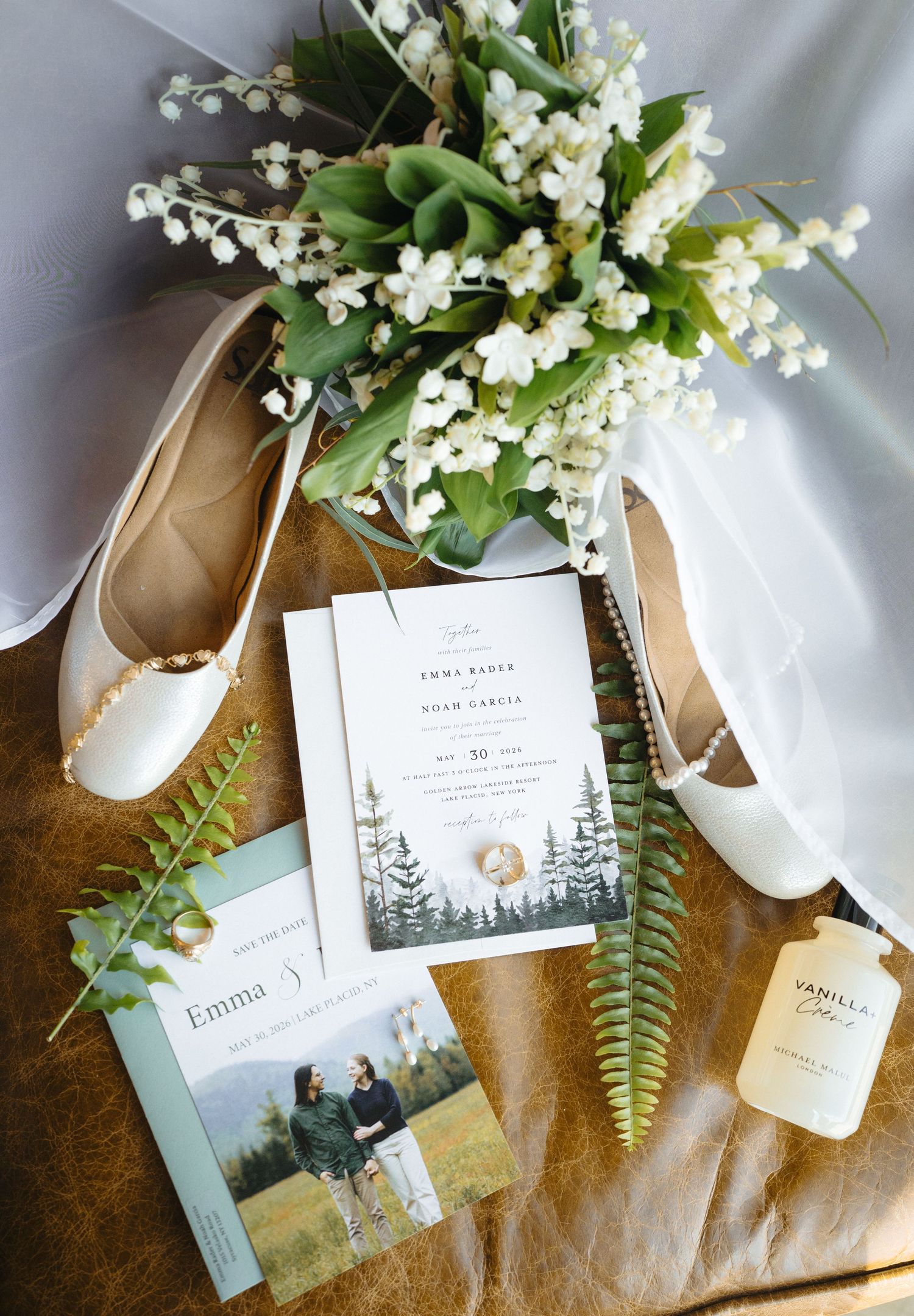 Wedding flat lay with white flats, lily of the valley bouquet, invitations, and vanilla fragrance on brown leather.