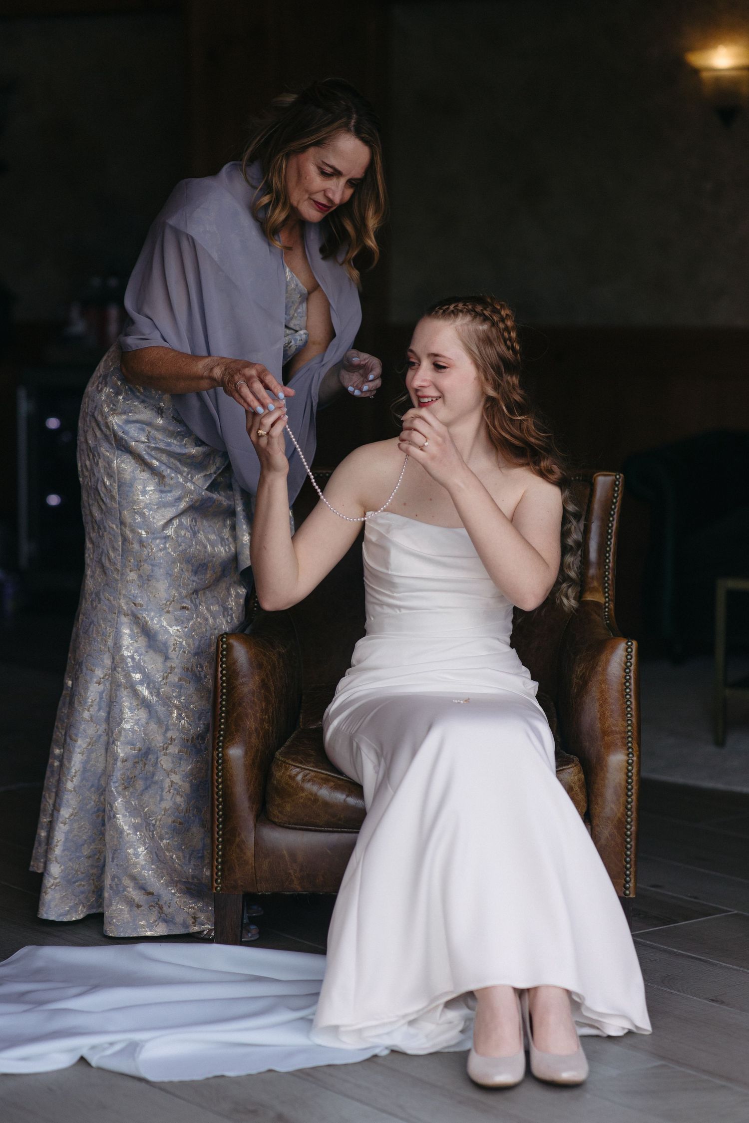 Bride in white gown laughing joyfully as woman in blue dress fastens bracelet in rustic venue.