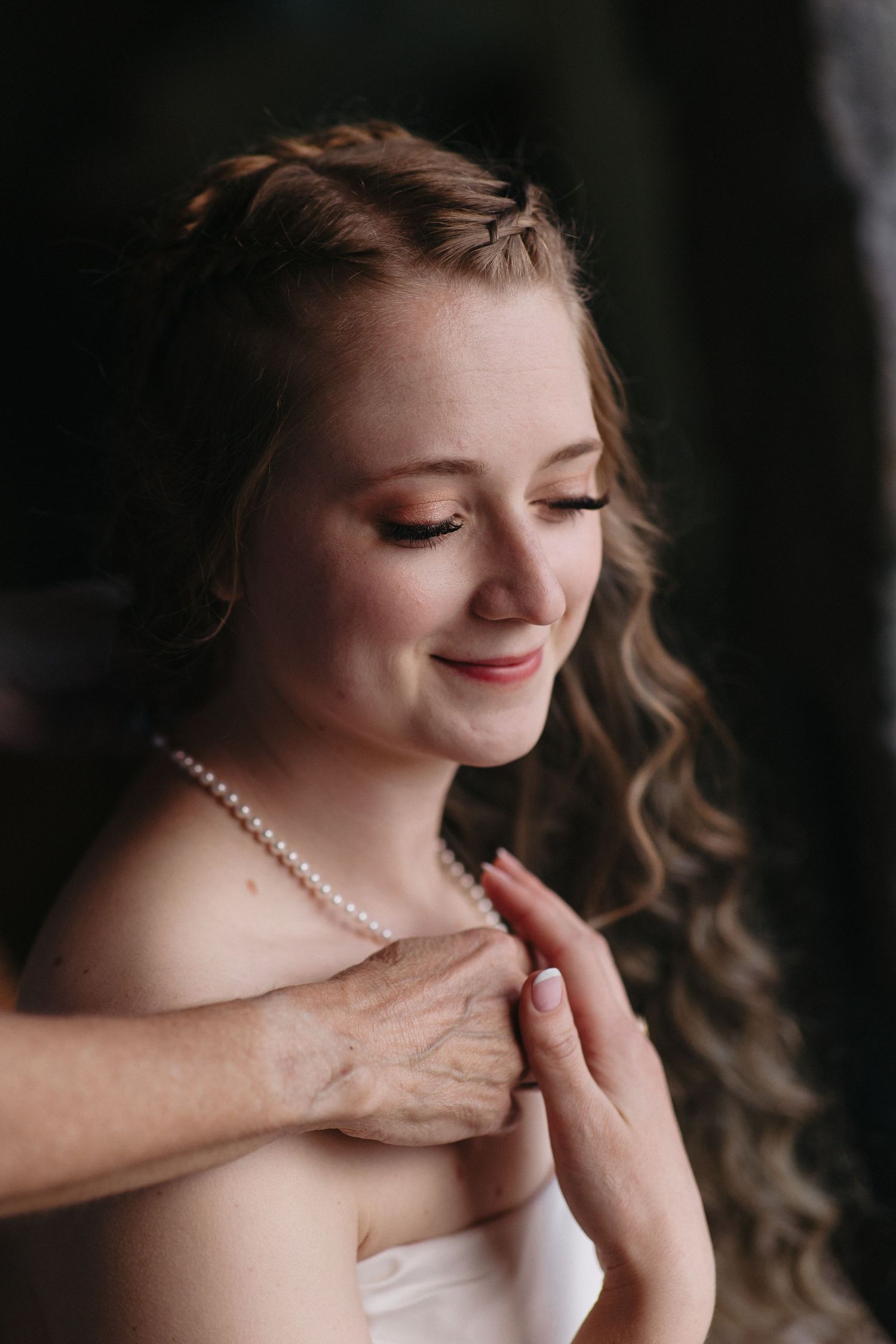 Bride smiling softly as pearl necklace is clasped, wearing elegant updo with braids and loose curls.
