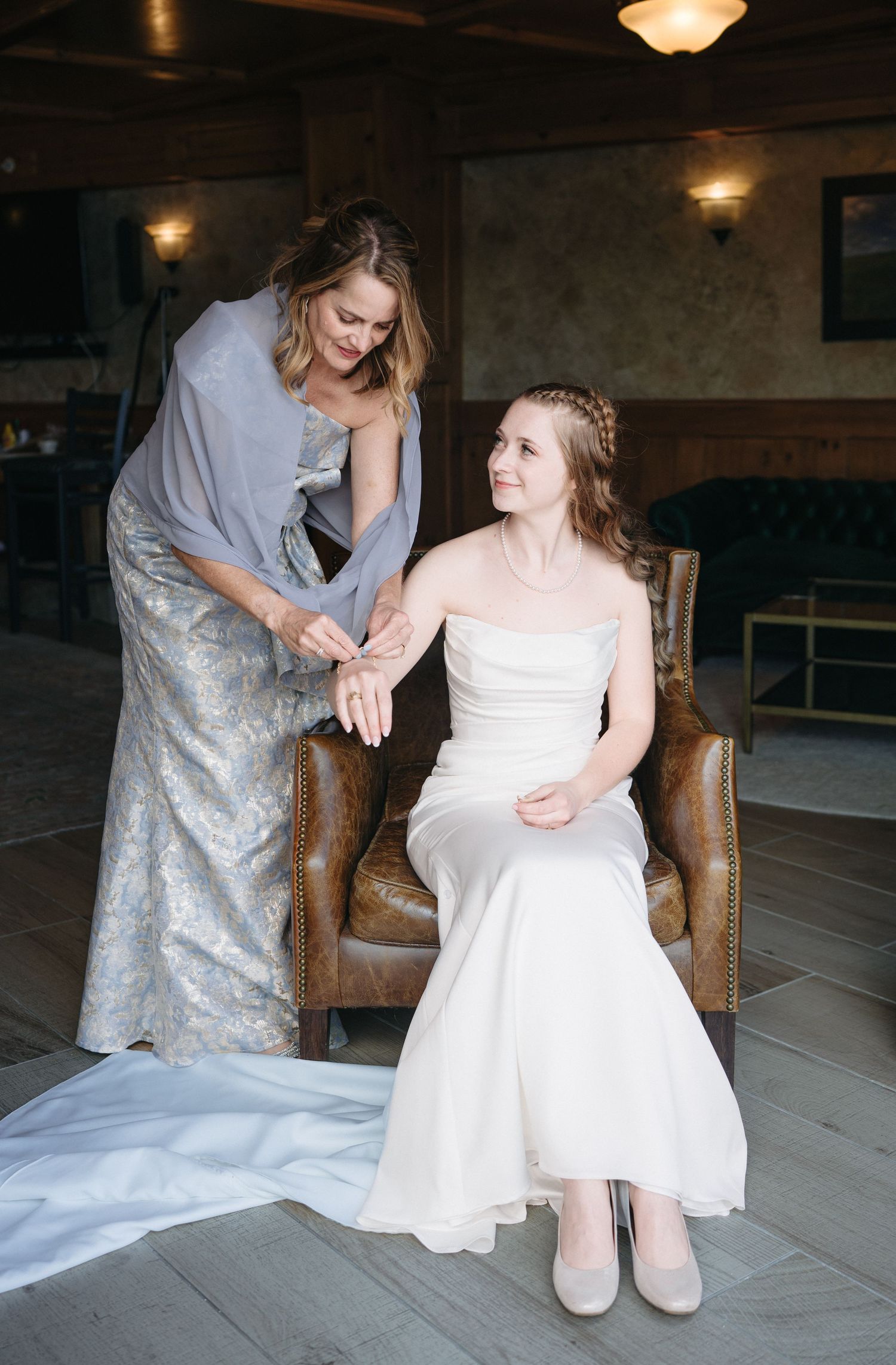 Mother helping bride in strapless white gown with jewelry while seated in leather chair indoors.