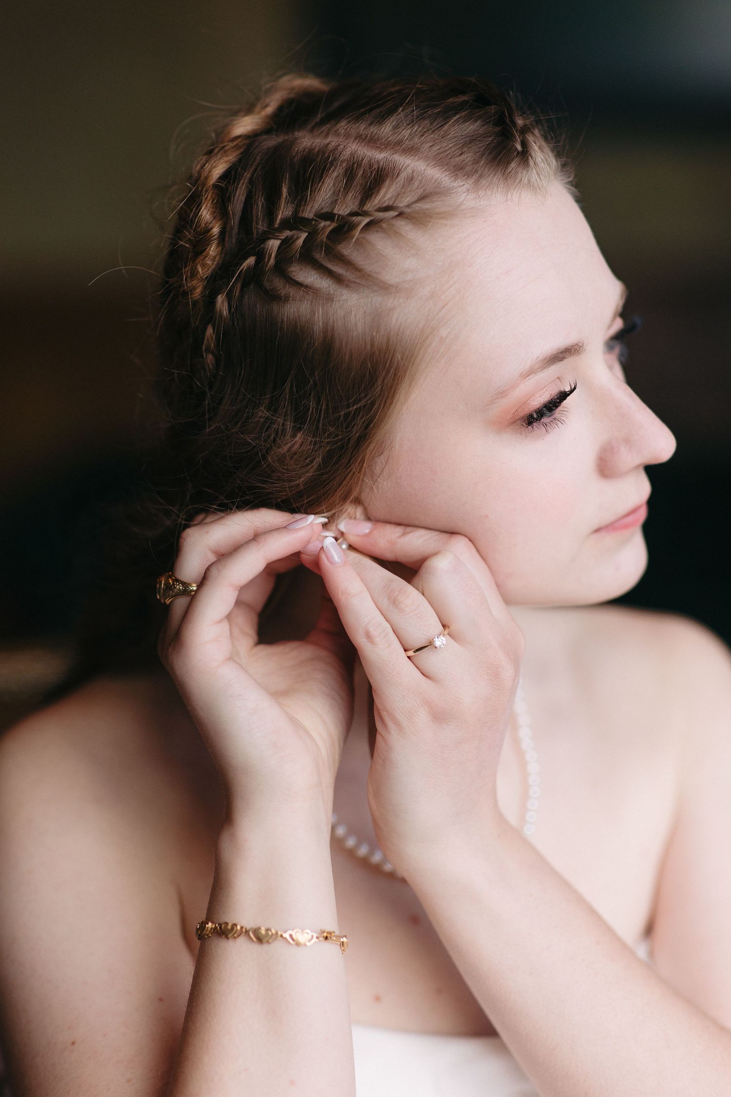 Close-up of bride with braided updo putting on earrings with pearl bracelet and gold jewelry.