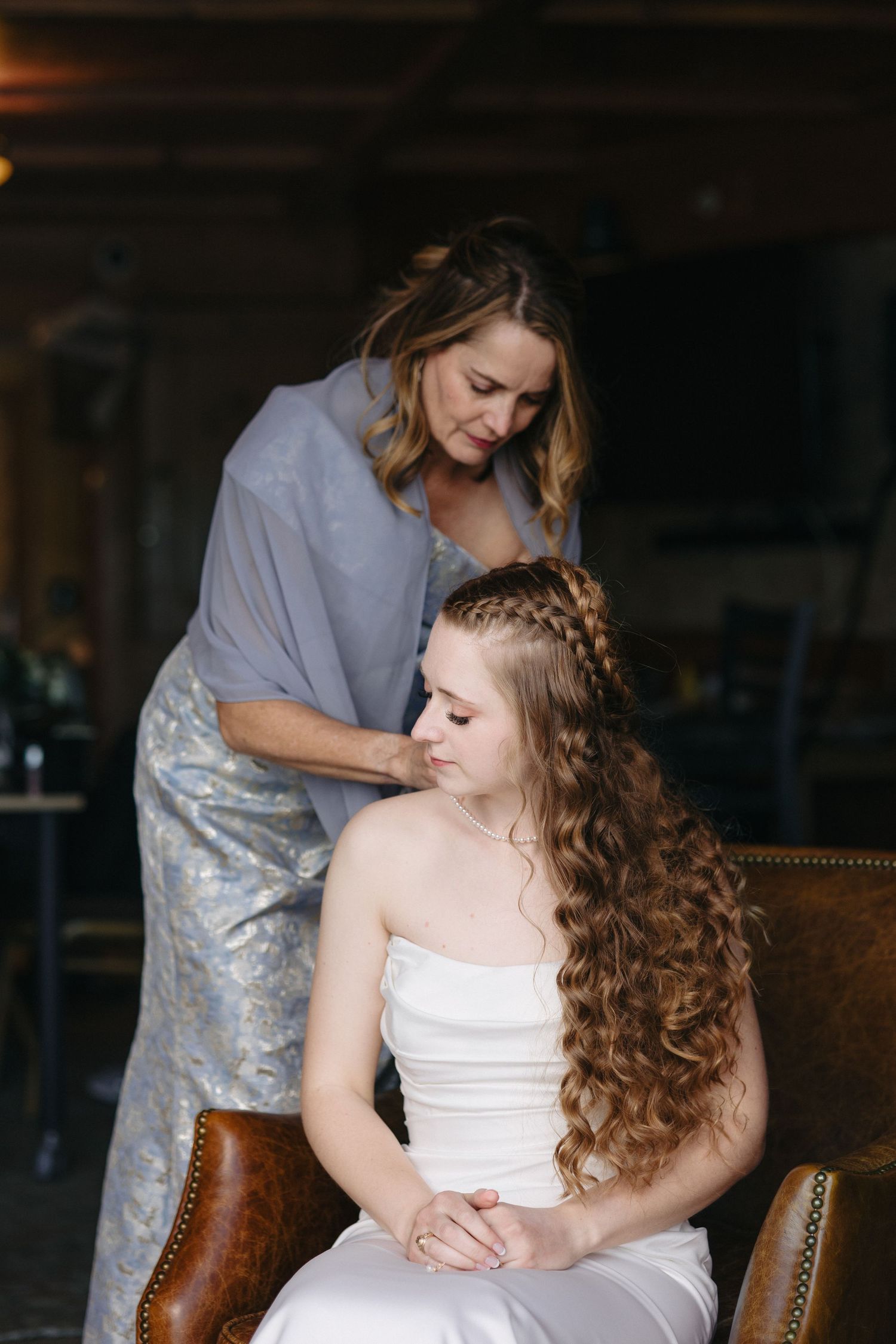 Mother helping bride with long curly hair and braid while seated in leather chair before wedding.