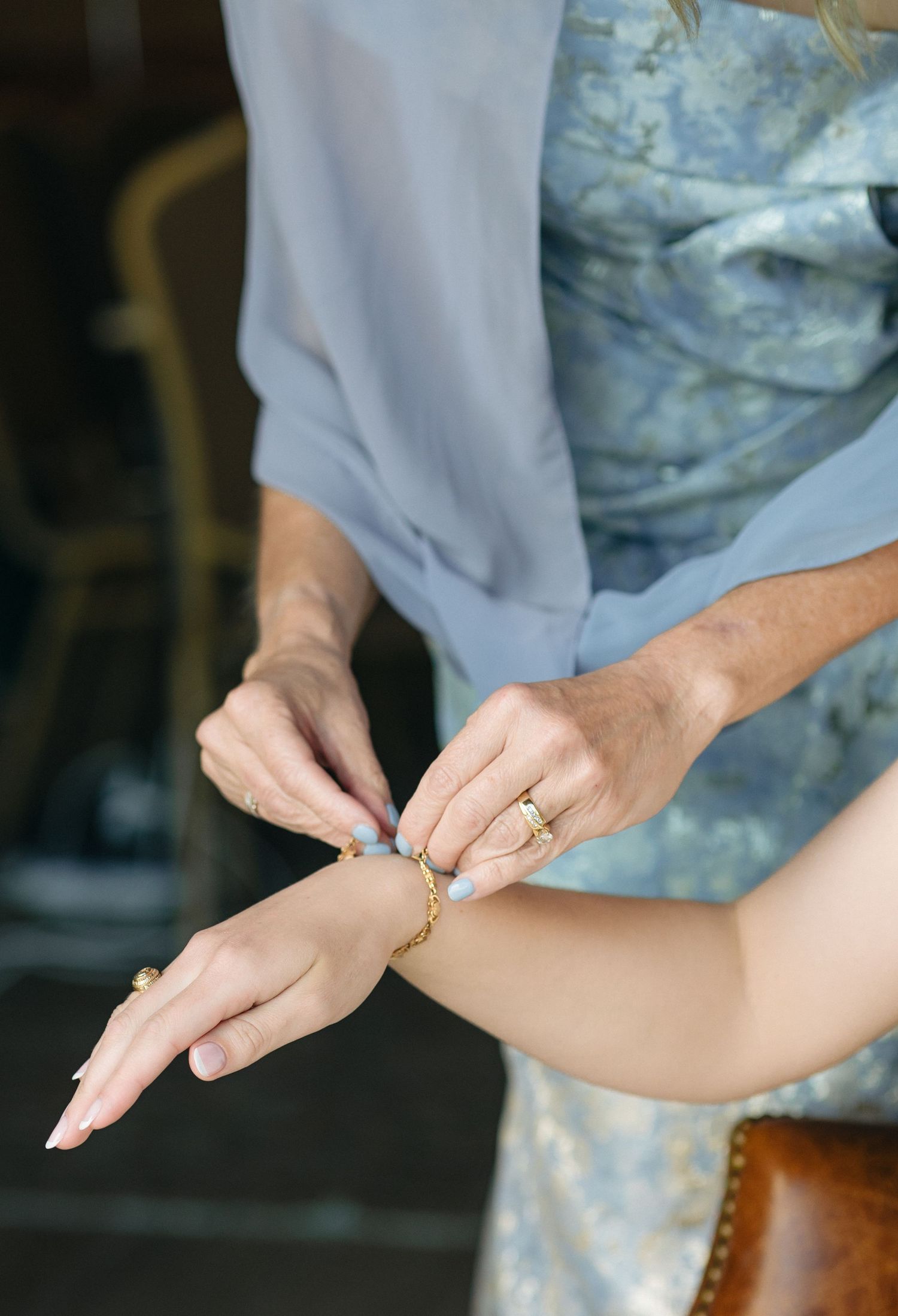 Mother fastening a gold bracelet on the wrist of a bride wearing a strapless white wedding gown.