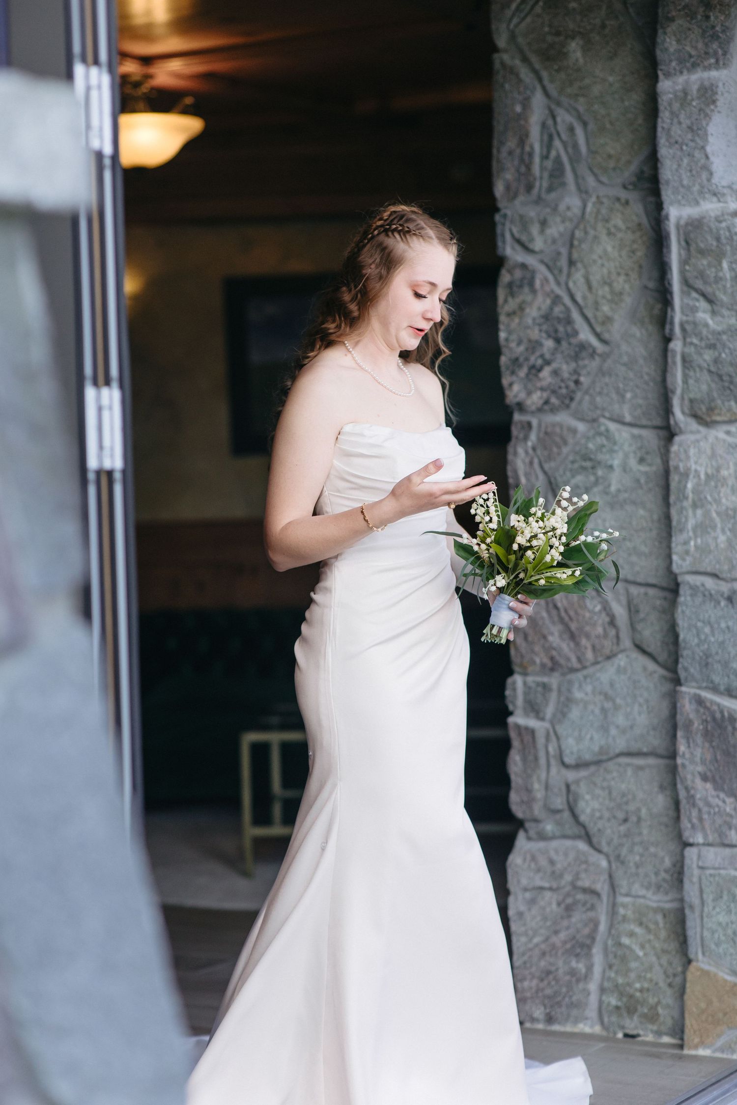 Bride in strapless white mermaid gown holding lily of the valley bouquet beside stone wall at venue.