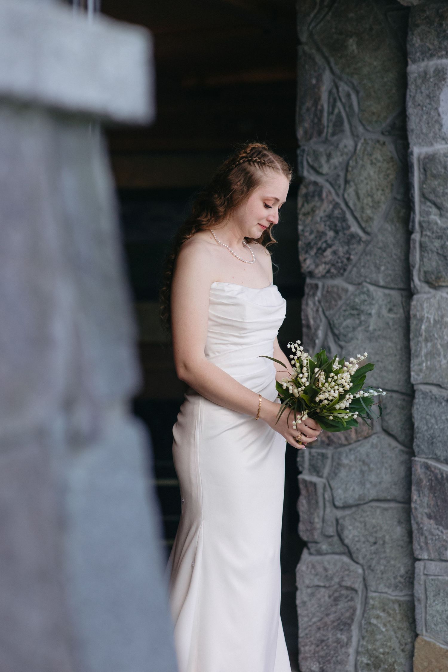 Bride in strapless white satin gown holding lily of the valley bouquet beside stone wall column.