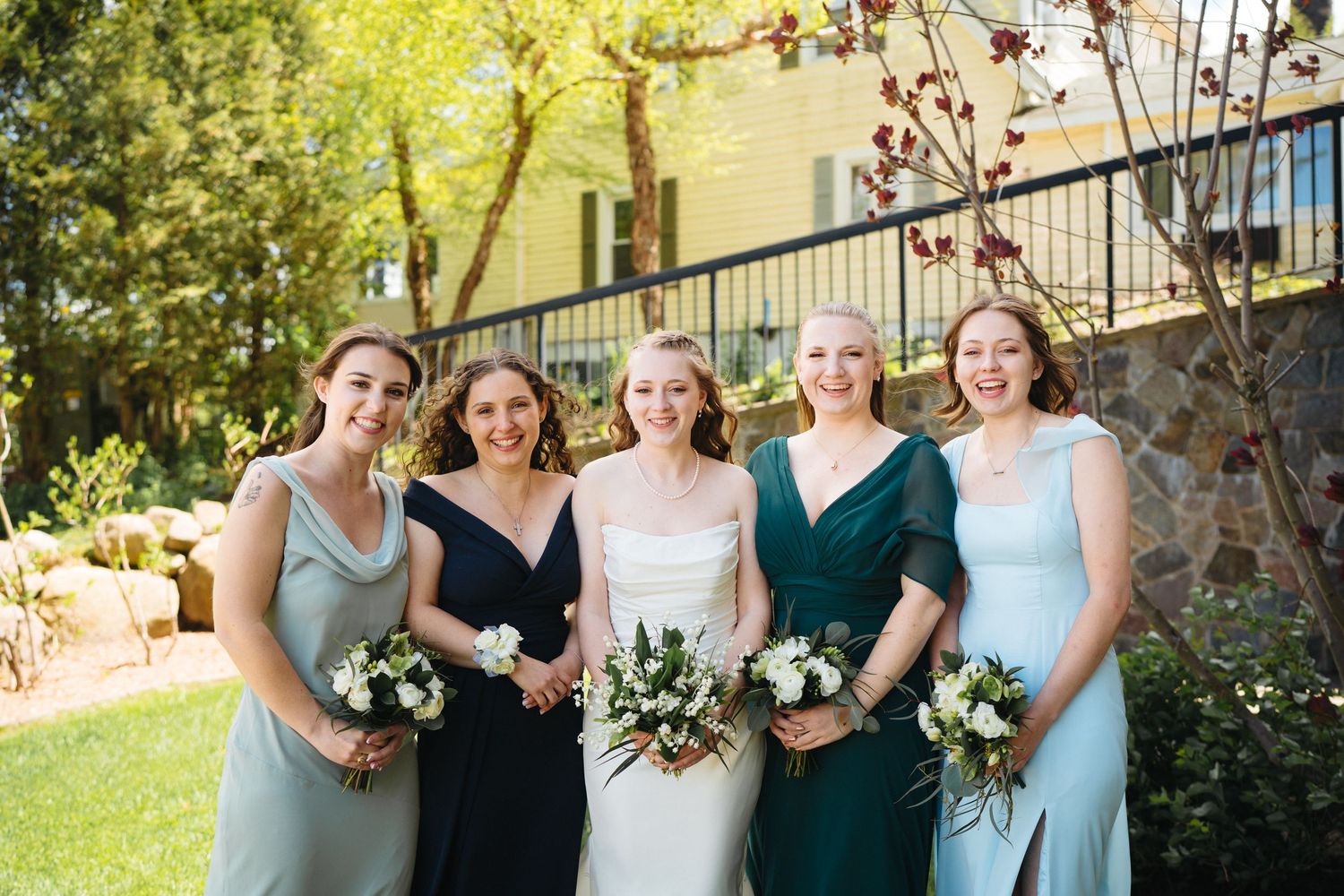 Bride with four bridesmaids in mismatched sage and dark green gowns holding white bouquets outdoors.