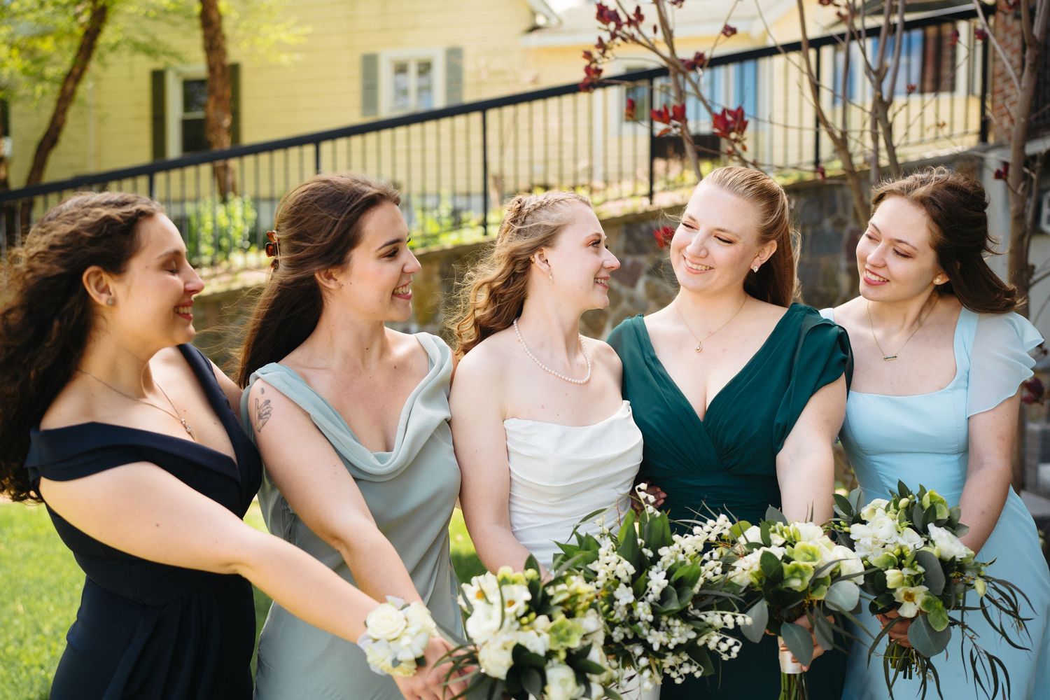Bride and four bridesmaids in mismatched blue and green dresses laughing together outdoors.