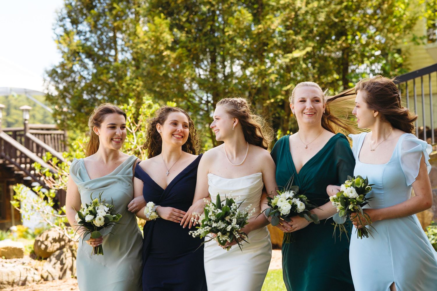 Bride and four bridesmaids in blue and green dresses walking outdoors holding white bouquets.
