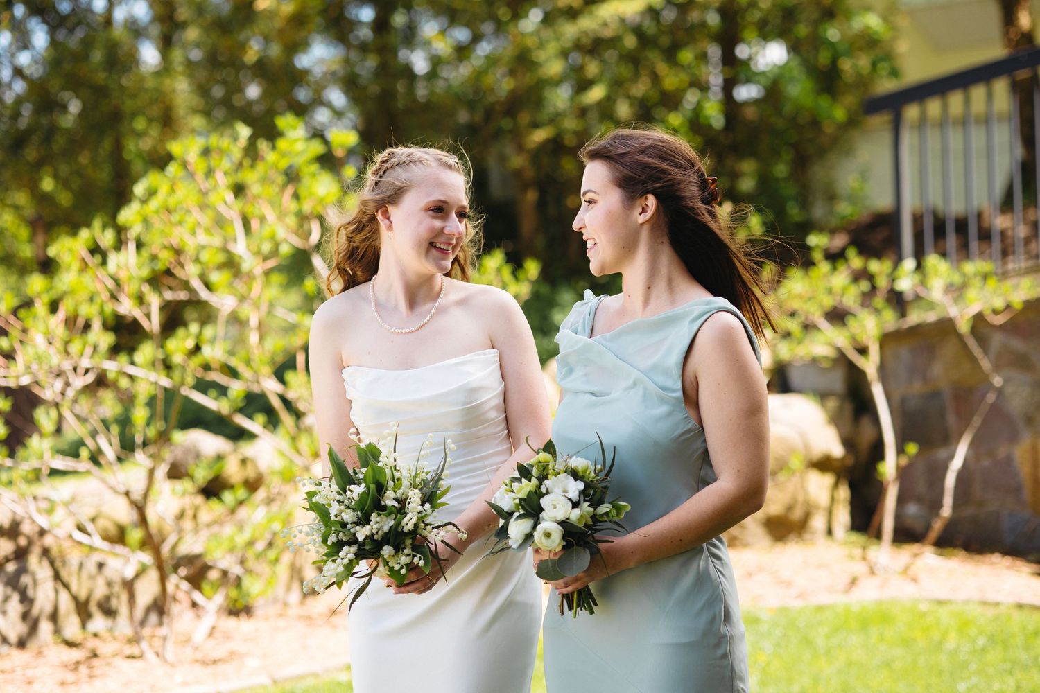 Smiling bride in white strapless gown and bridesmaid in mint dress holding bouquets in sunny garden.