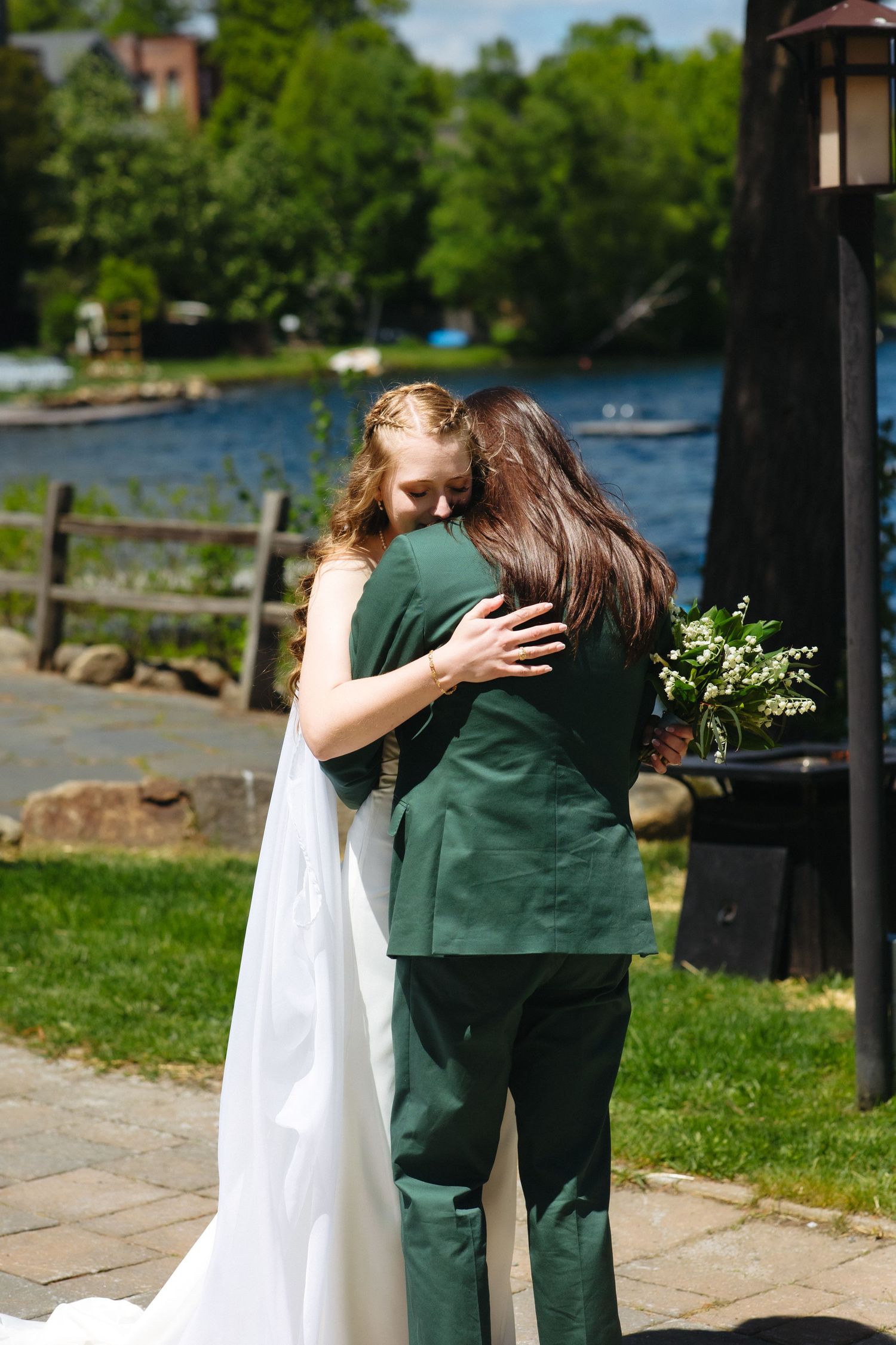 Bride in white gown and groom in green suit share an emotional hug outdoors by a lakeside venue.