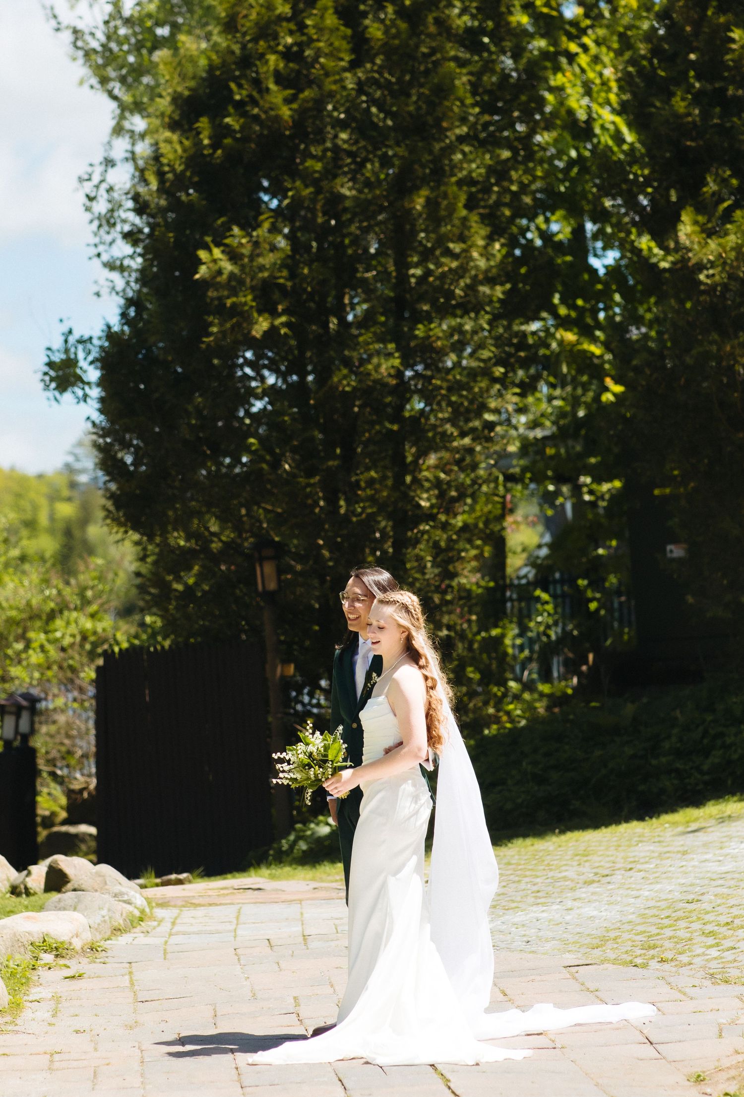 Bride and groom walk together on a sunlit stone path surrounded by lush green trees.