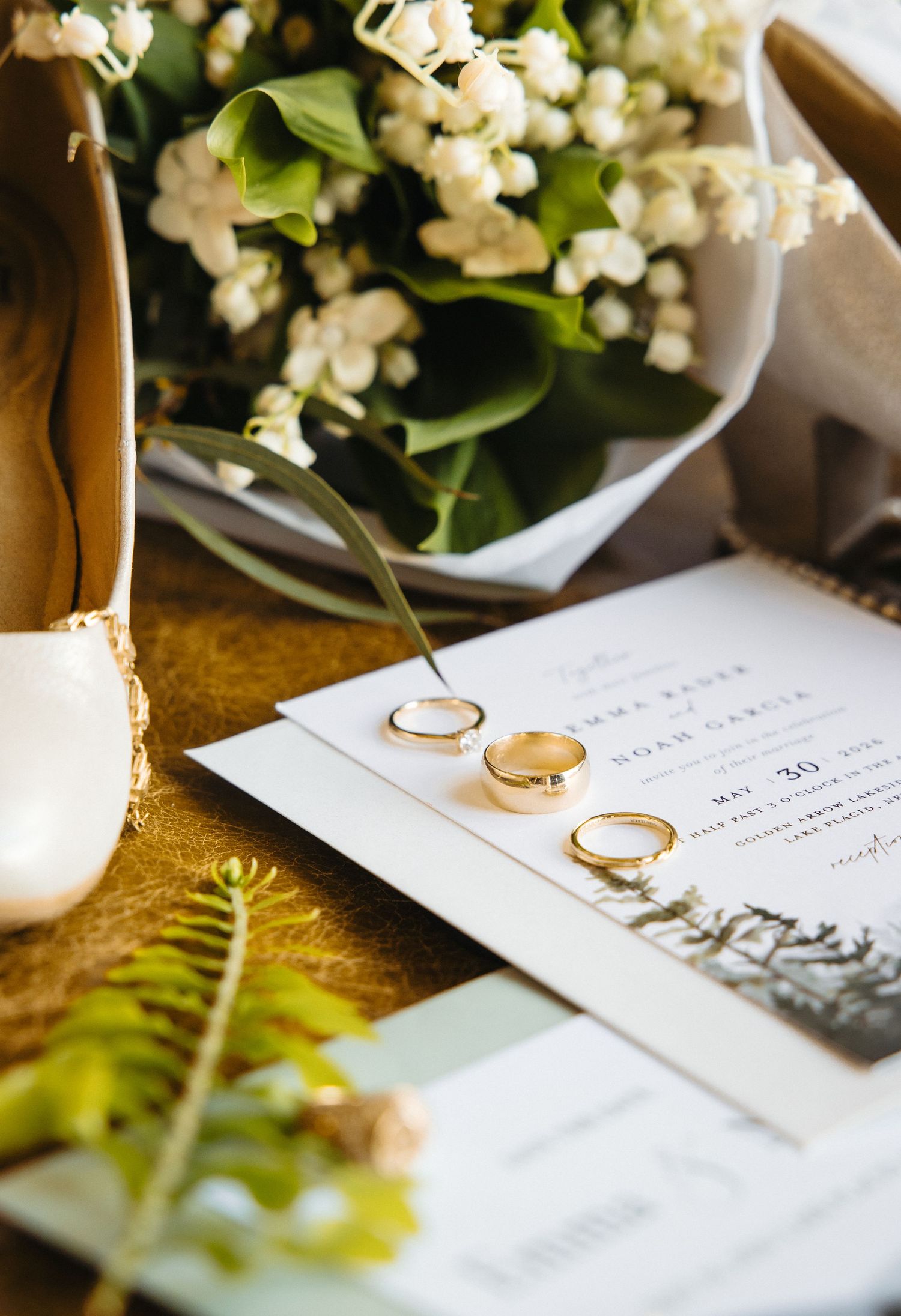 Gold wedding bands displayed on an invitation suite beside white floral bouquet, bridal shoe, and fern leaf.
