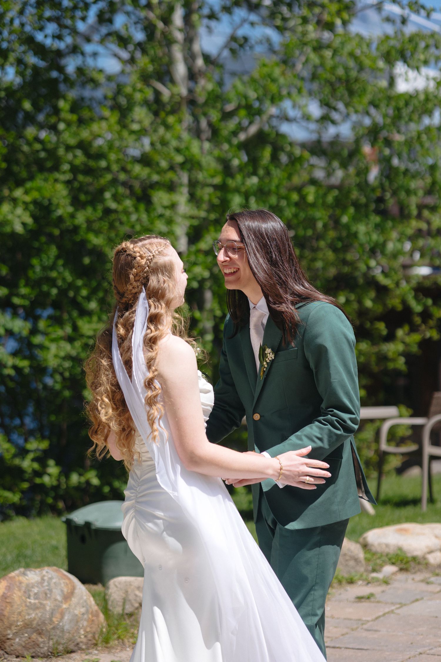 Smiling bride and groom in green suit share a tender moment together in a sunny outdoor garden setting.