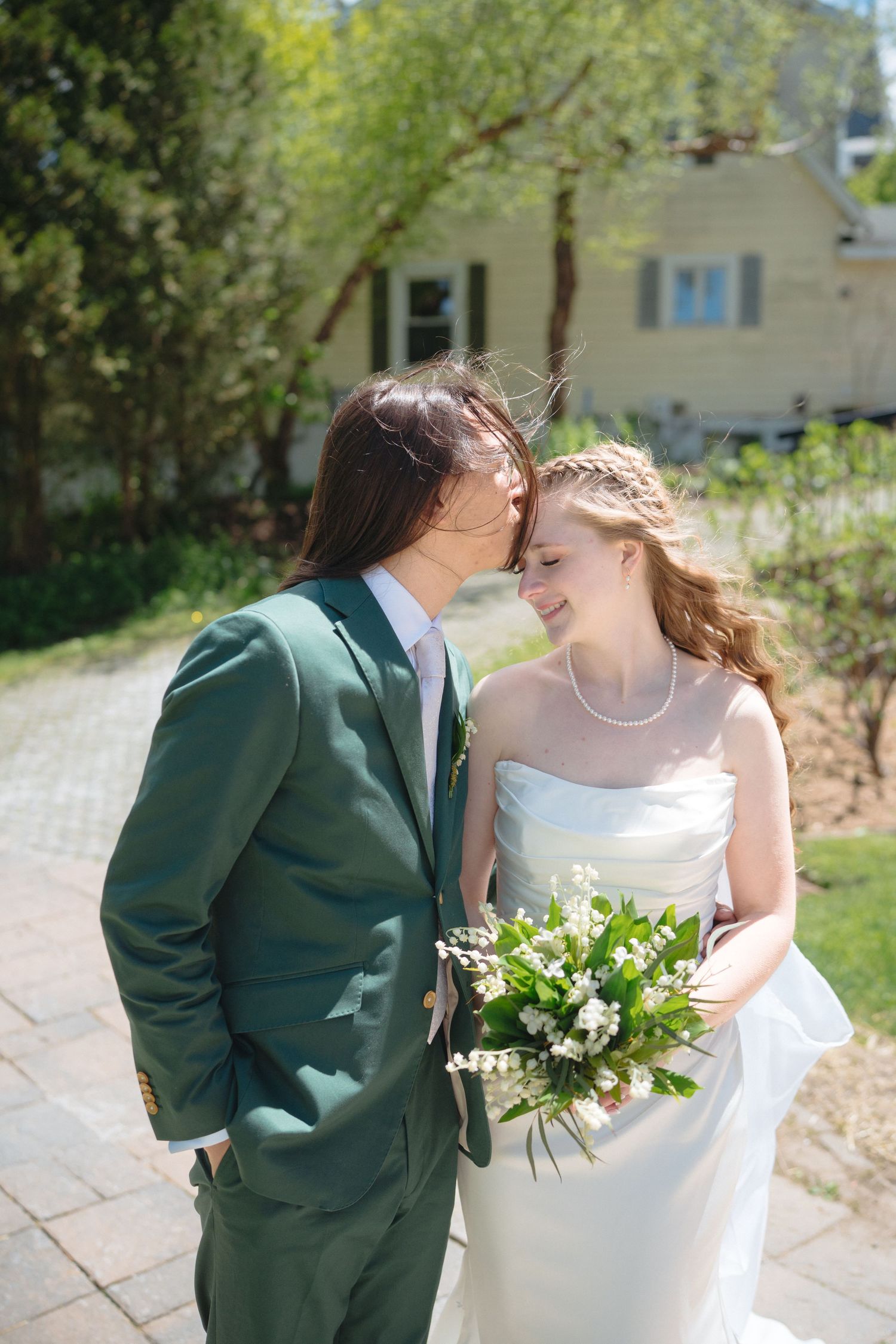 Bride holding white bouquet and groom in green suit share a tender moment in a sunny garden setting.