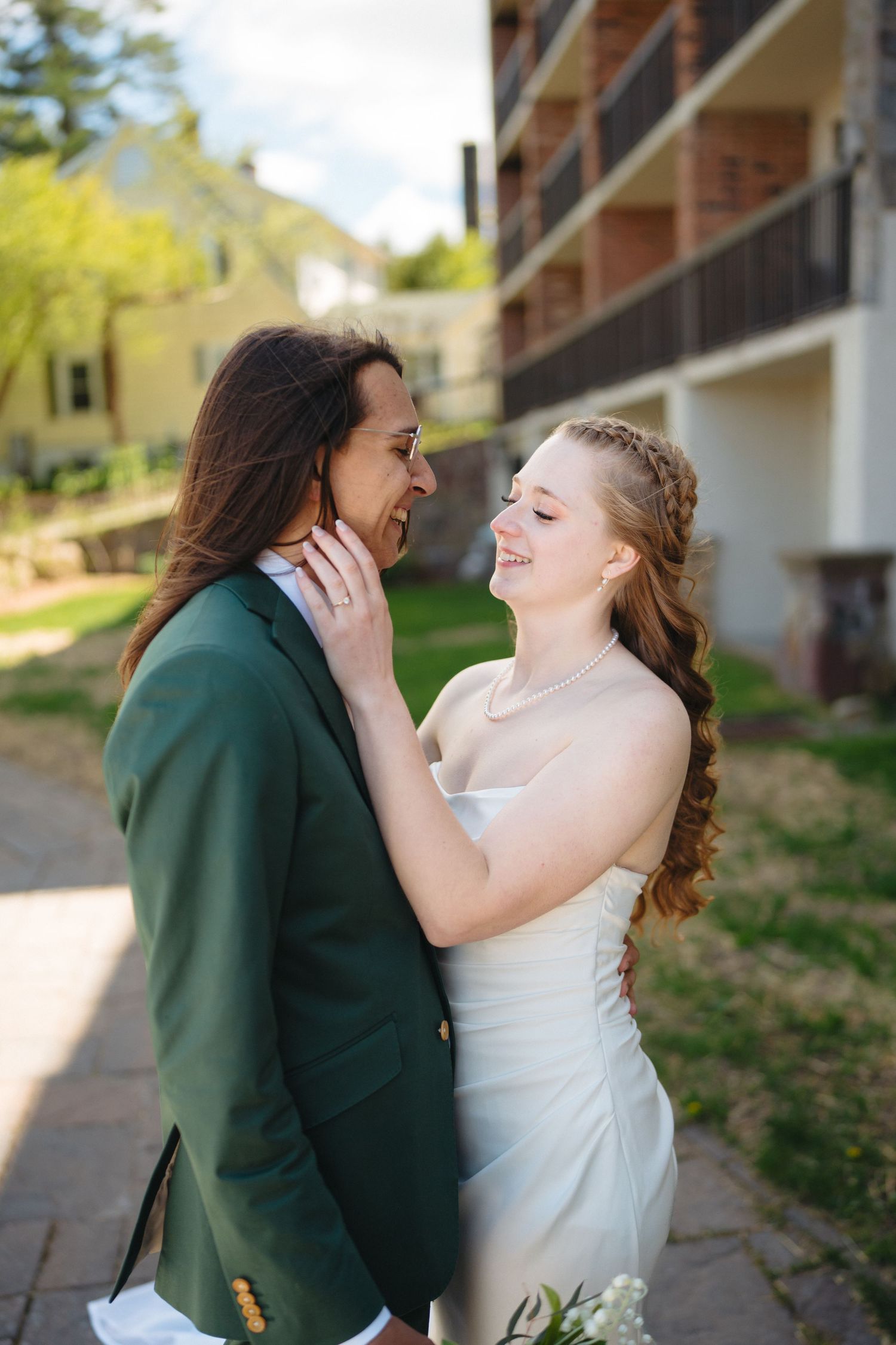 Bride in white gown smiles at groom in green suit while touching his face outdoors.