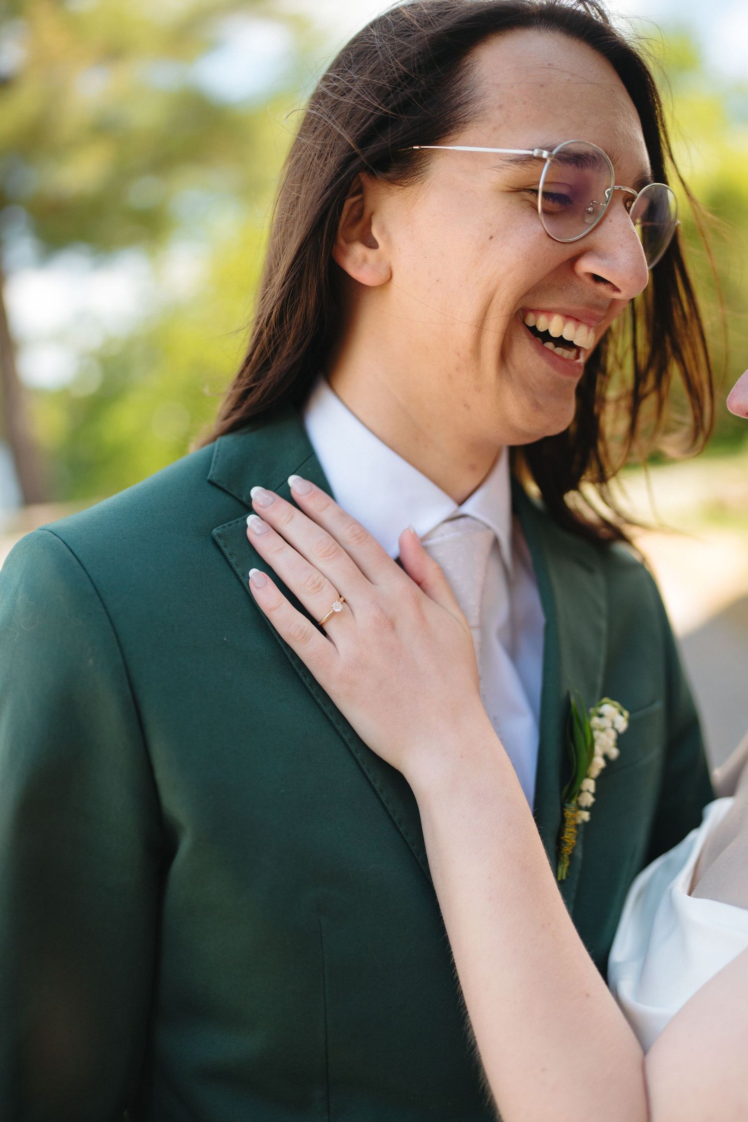 Laughing groom in green suit with glasses as bride places hand on his chest outdoors.