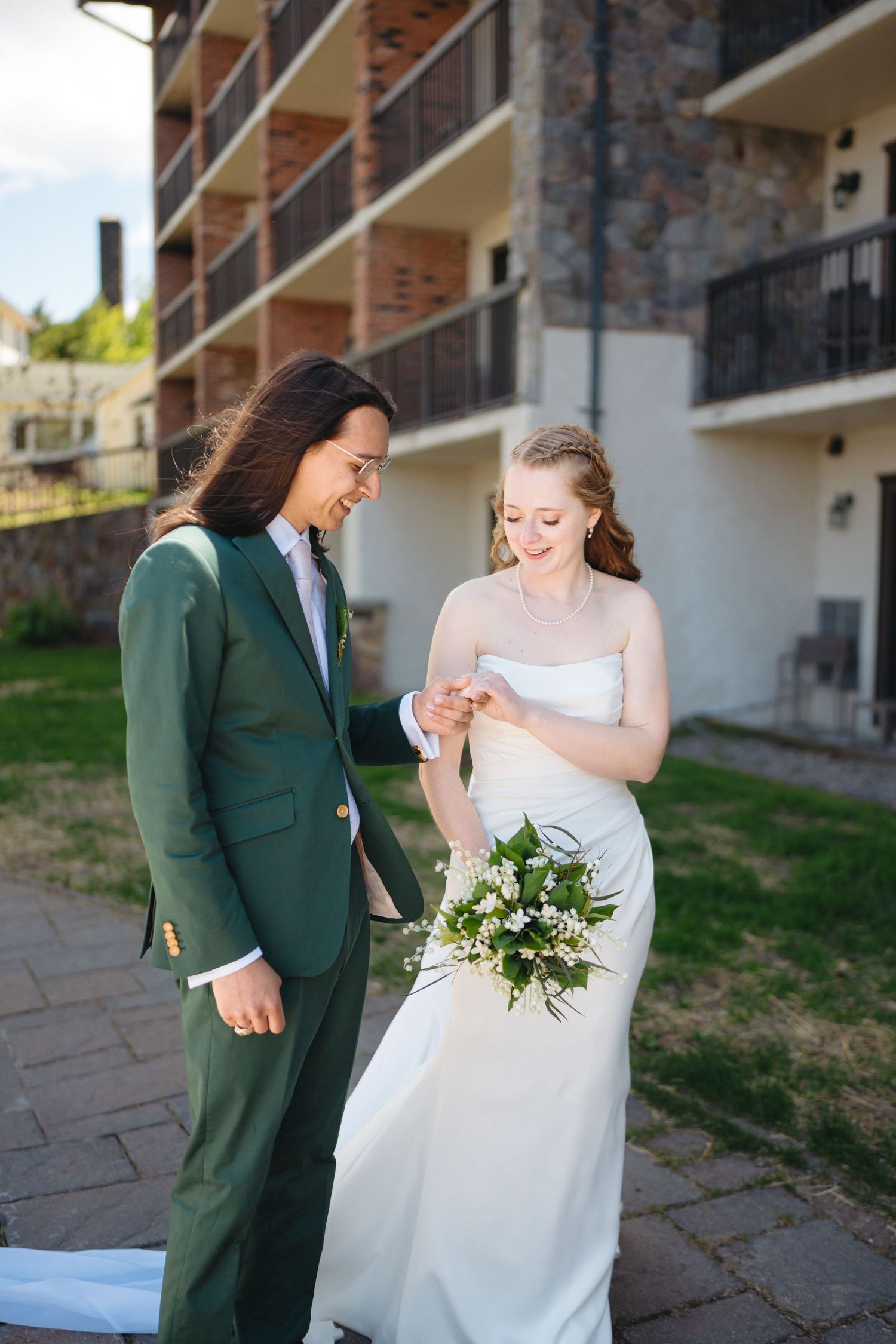 Groom in green suit places wedding ring on brides finger outdoors near a stone building.
