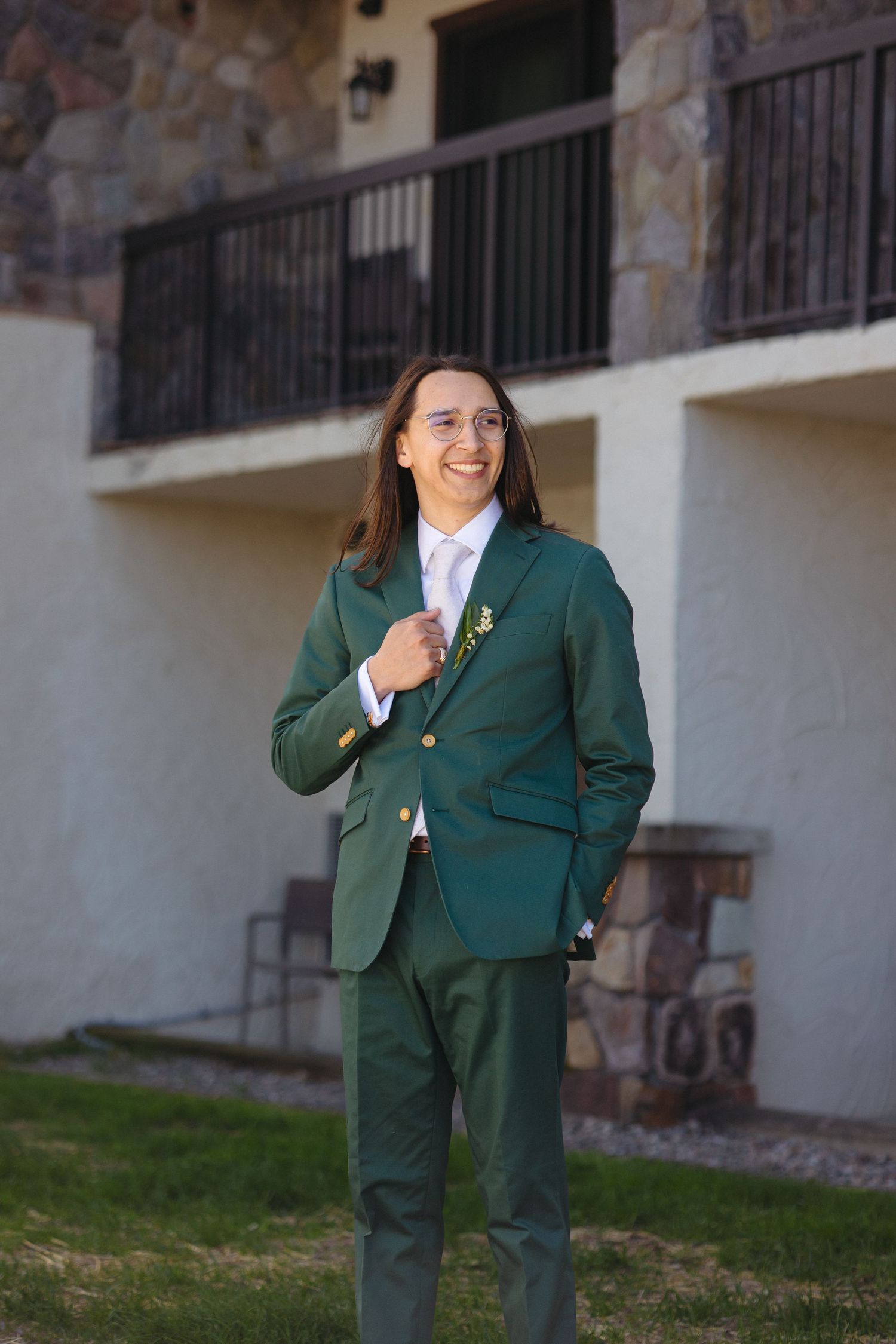 Groom smiling confidently in a dark green suit with boutonniere outside a stone building.