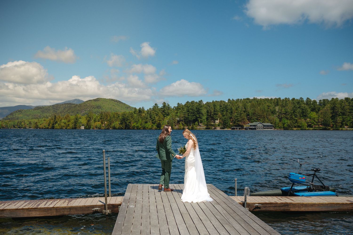 Bride and groom hold hands on a wooden dock overlooking a stunning blue mountain lake on their wedding day.