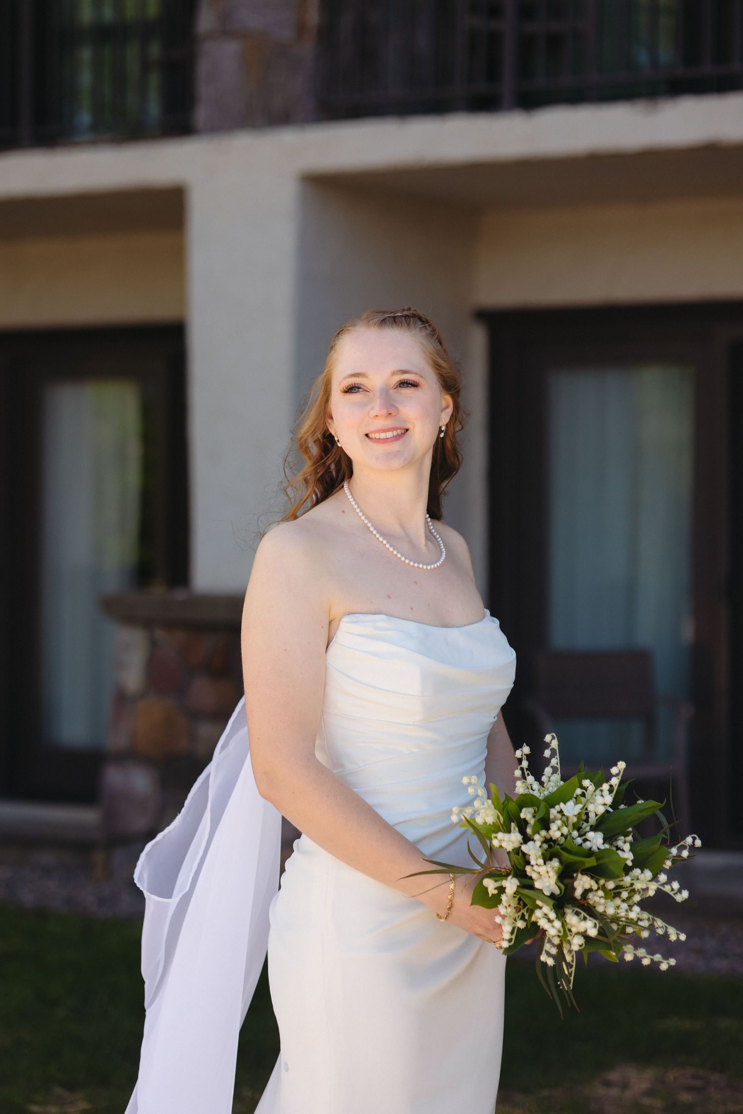 Smiling bride in a white strapless gown with pearl necklace holding a lily of the valley bouquet outdoors.