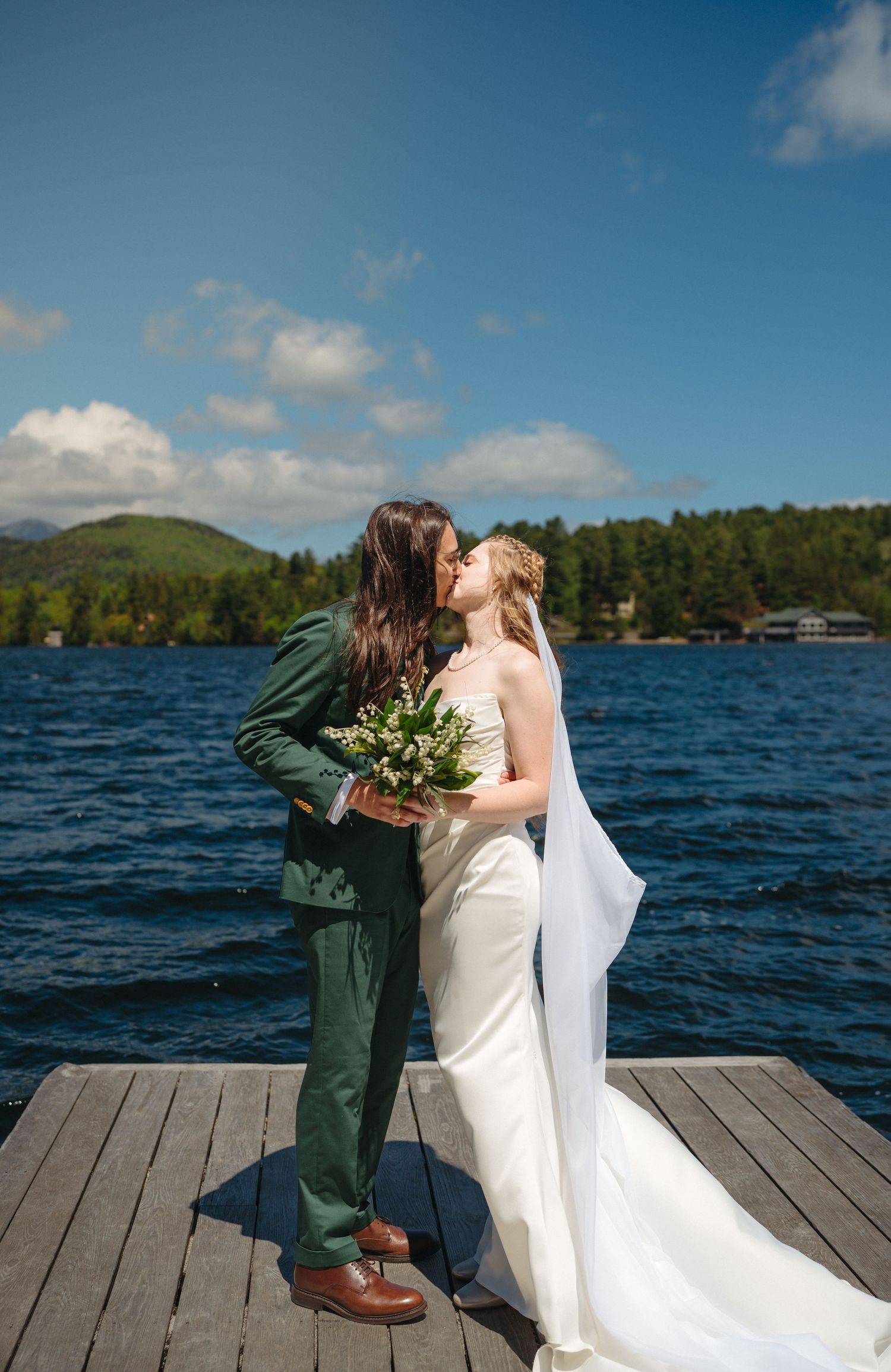 Newlyweds kissing on a lakeside dock, bride holding a green bouquet in a white gown with a long veil.