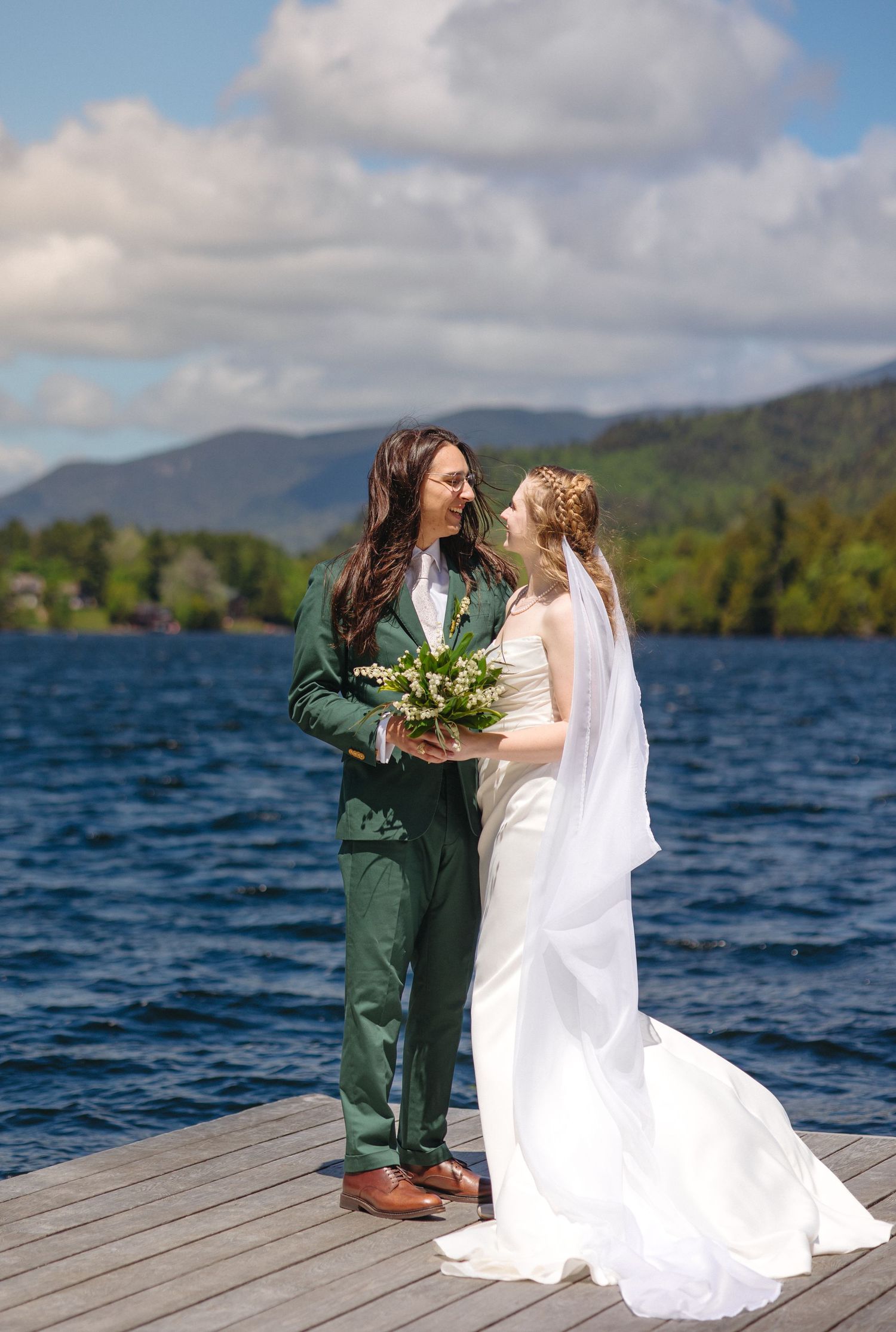Bride and groom stand on a lakeside dock holding a white bouquet with scenic mountains in the background.