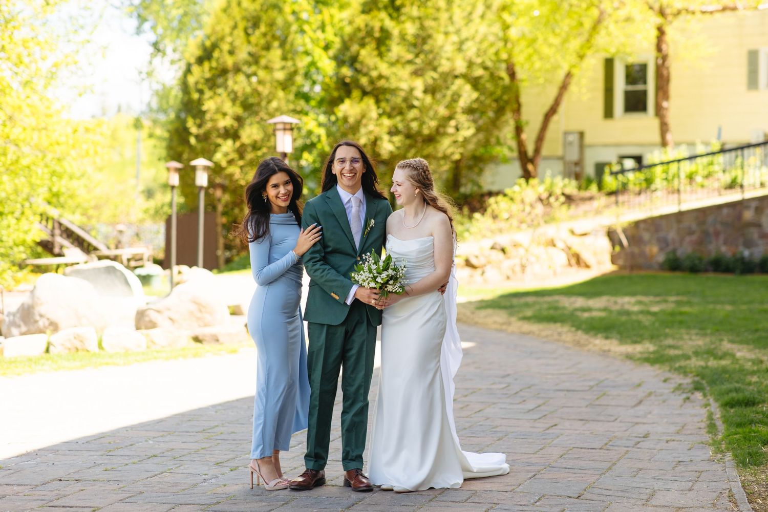 Bride, groom in green suit, and guest posing together outdoors on sunny wedding day.