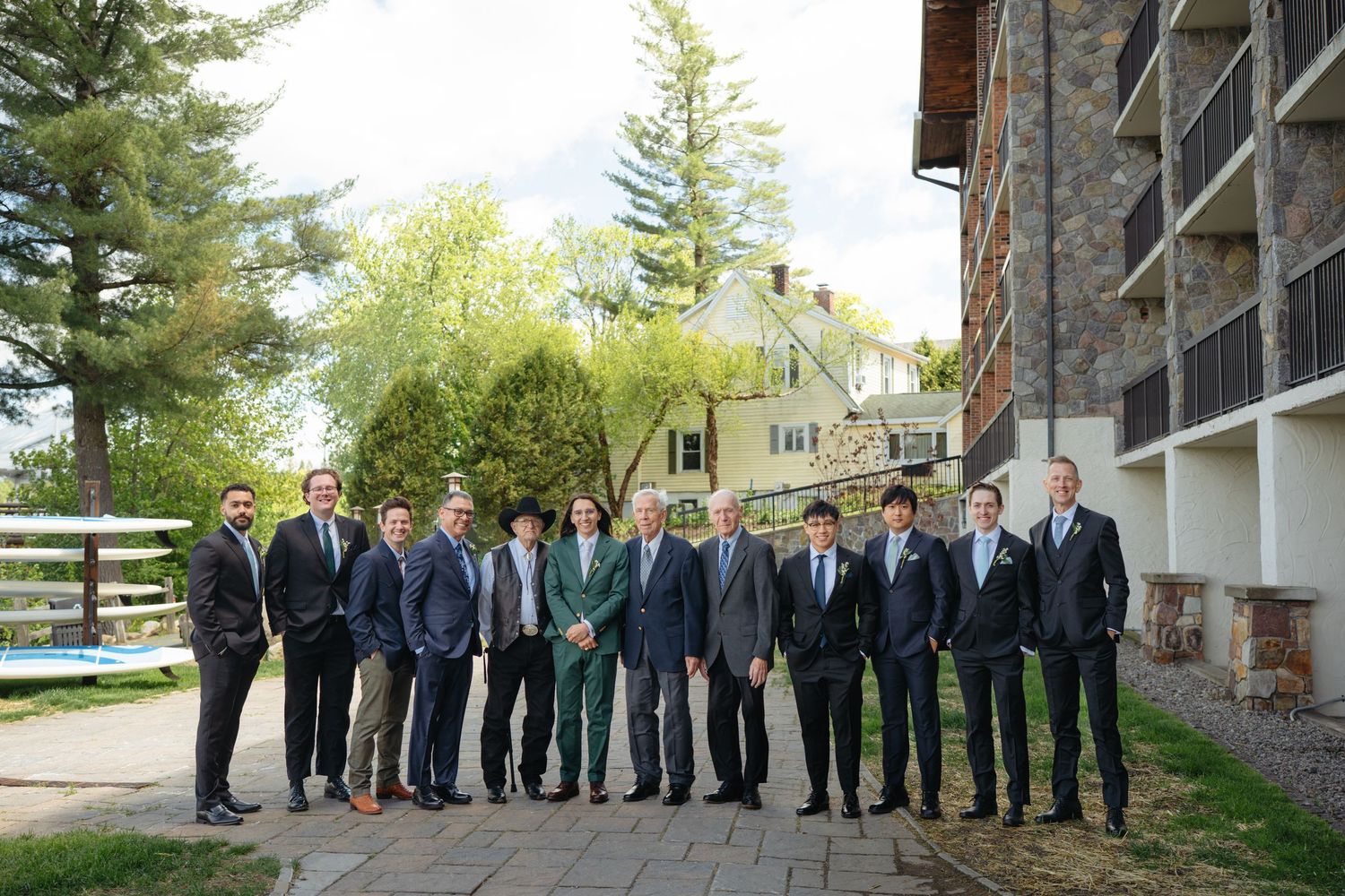 Groom and groomsmen in dark suits posing outdoors near stone building surrounded by spring greenery.