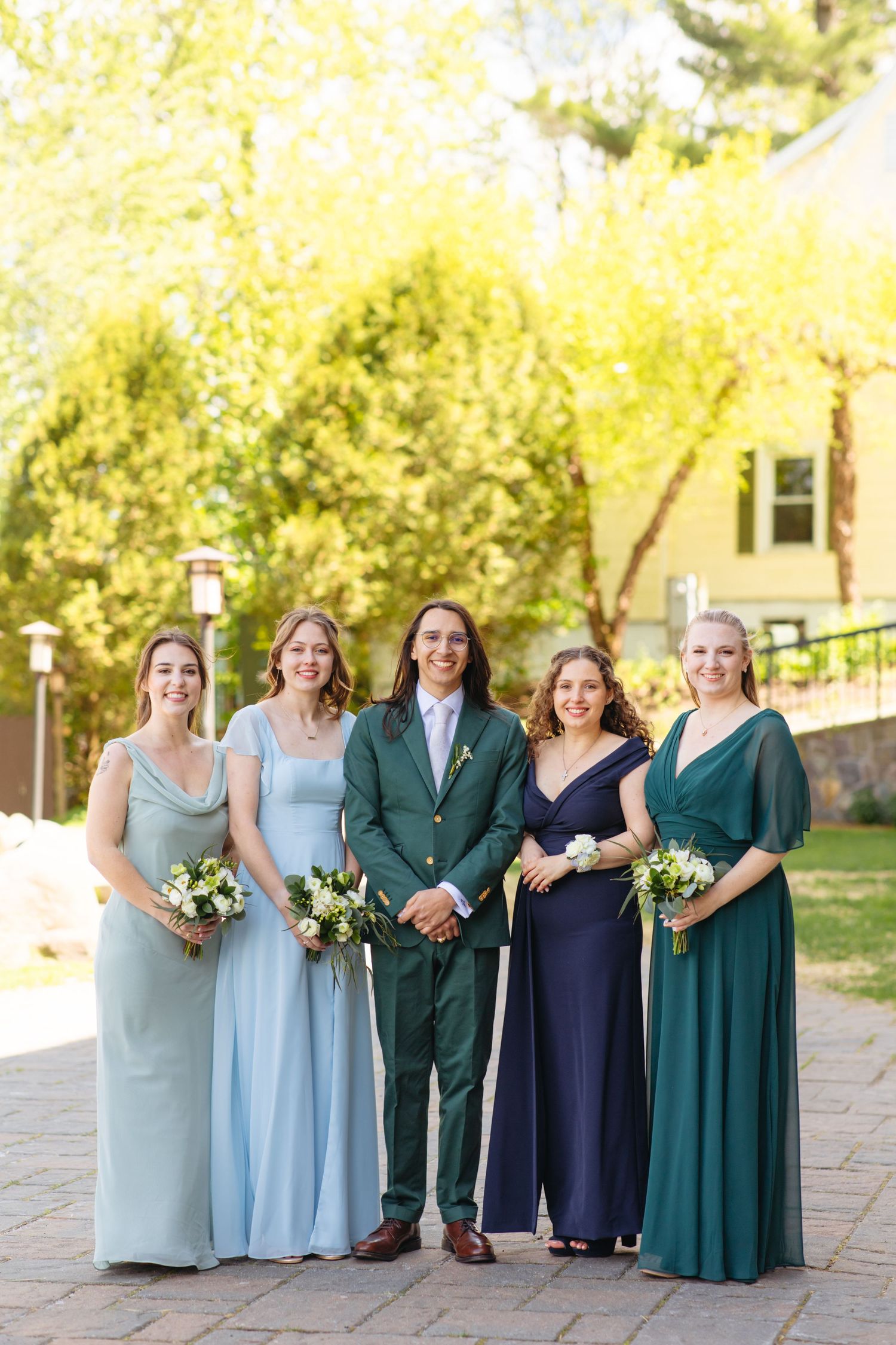 Wedding party posing outdoors in blue, navy, and green gowns with one person in a dark green suit.
