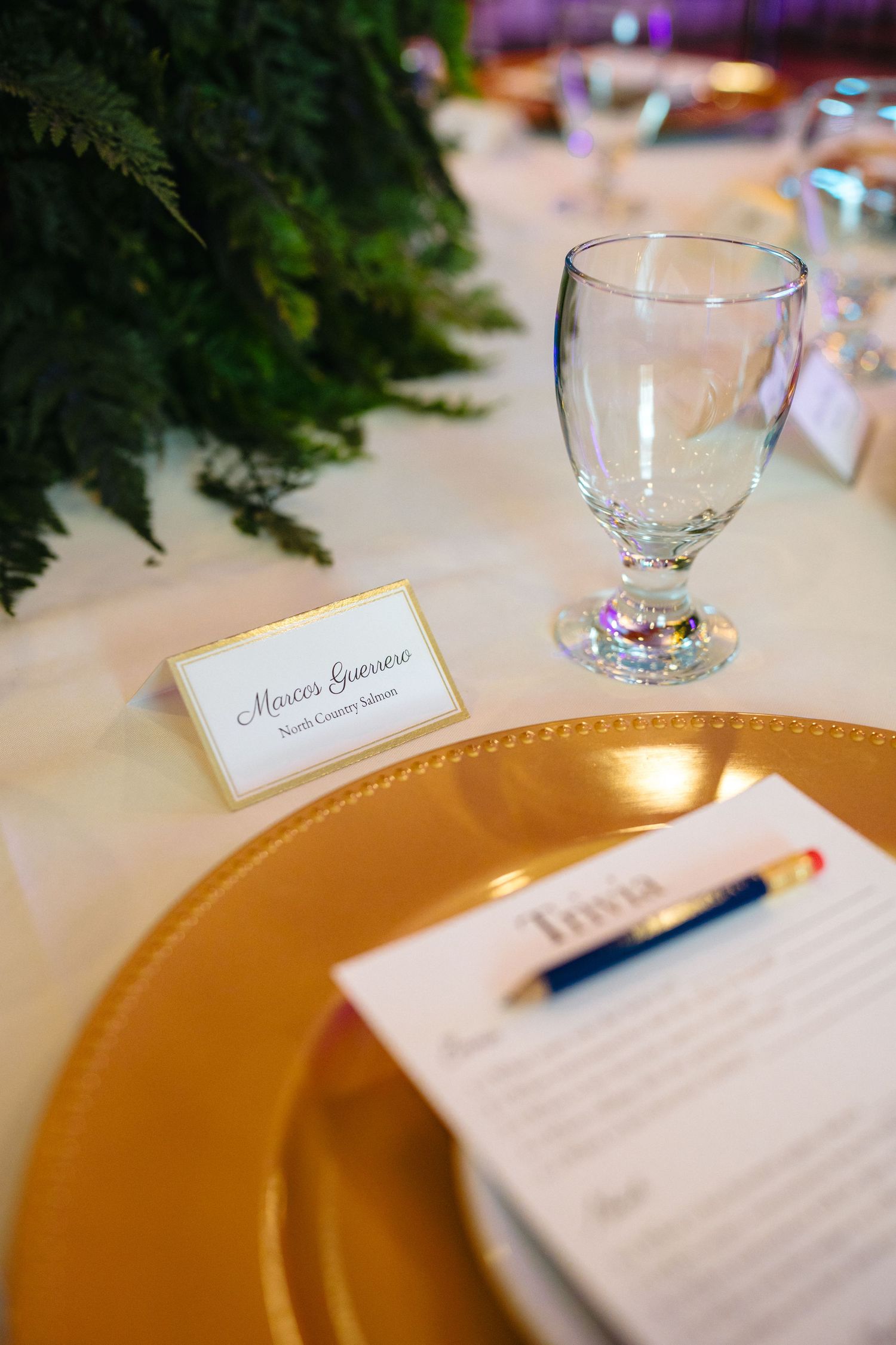 Gold charger plate with trivia sheet, pen, and place card for Marcus Genovese at an elegant wedding reception table.