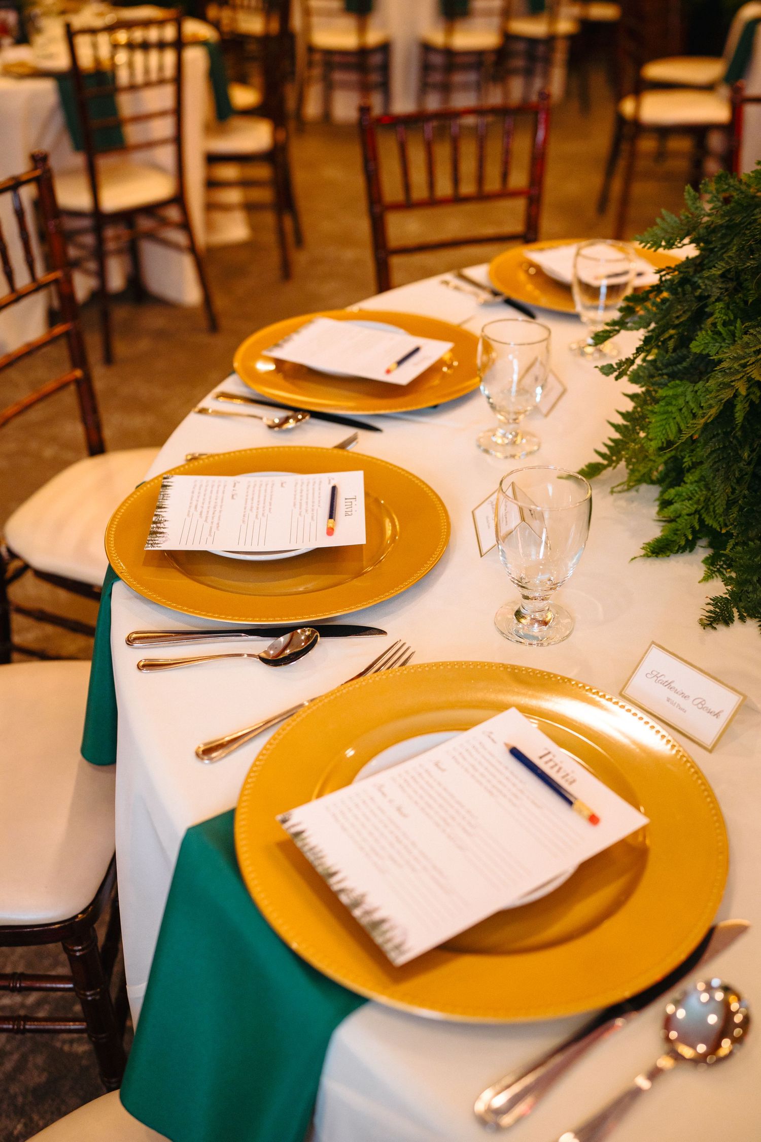 Elegant wedding reception table set with gold charger plates, teal napkins, crystal glasses, and greenery centerpiece.