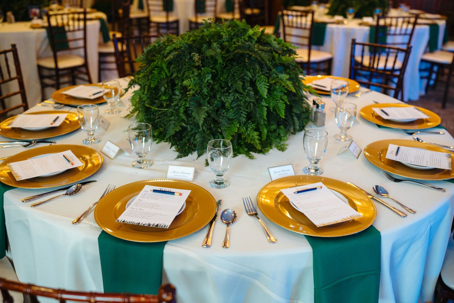 Elegant wedding reception table with gold charger plates, teal linen, and a lush green floral centerpiece.