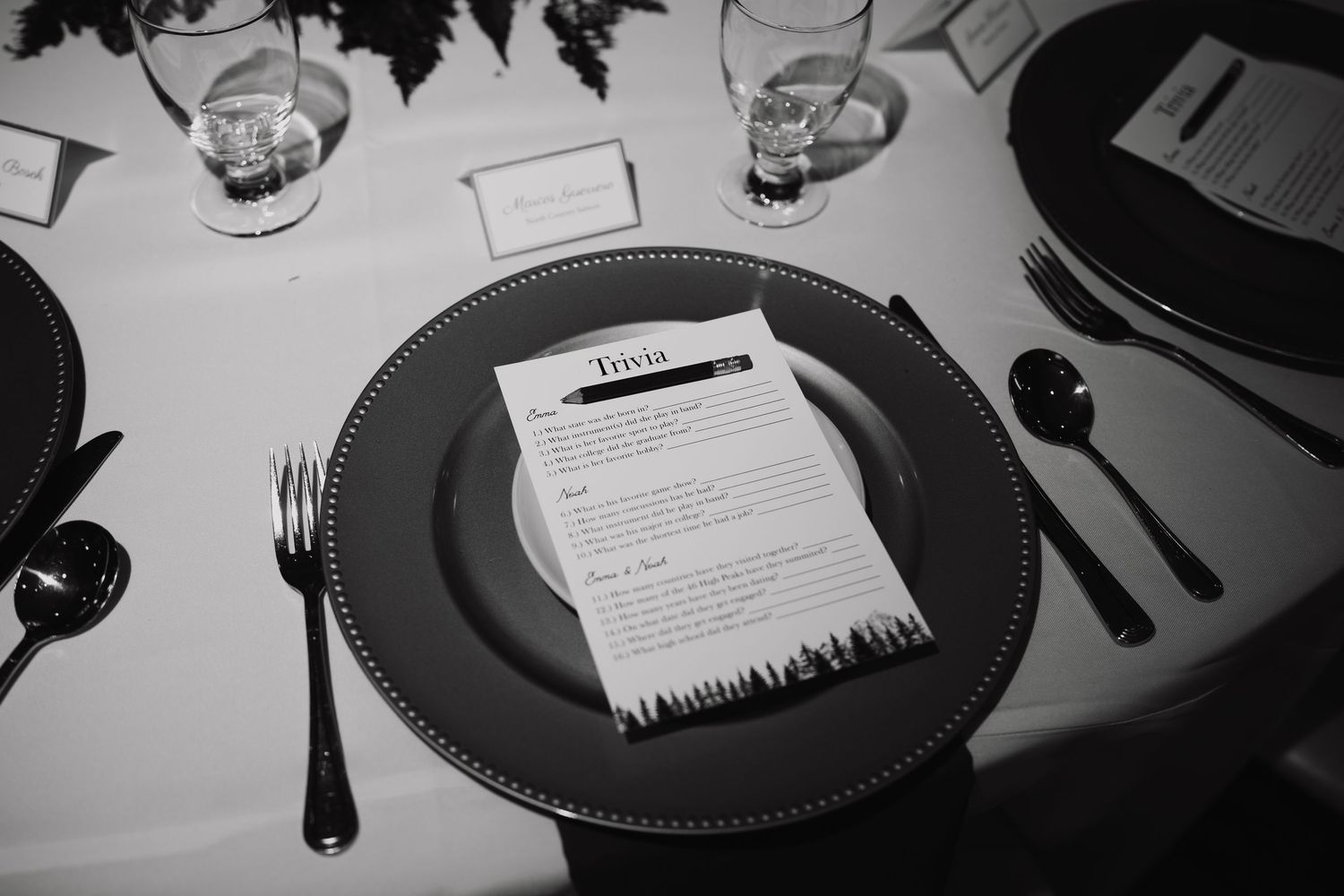 Elegant black and white wedding place setting with charger plate, trivia card, and formal cutlery on white linen.