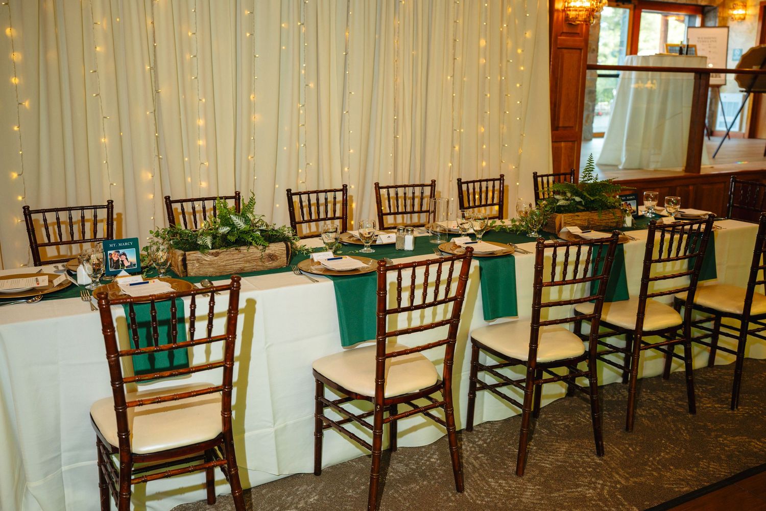Wedding reception head table decorated with teal runners, greenery centerpieces, and chiavari chairs with fairy lights.