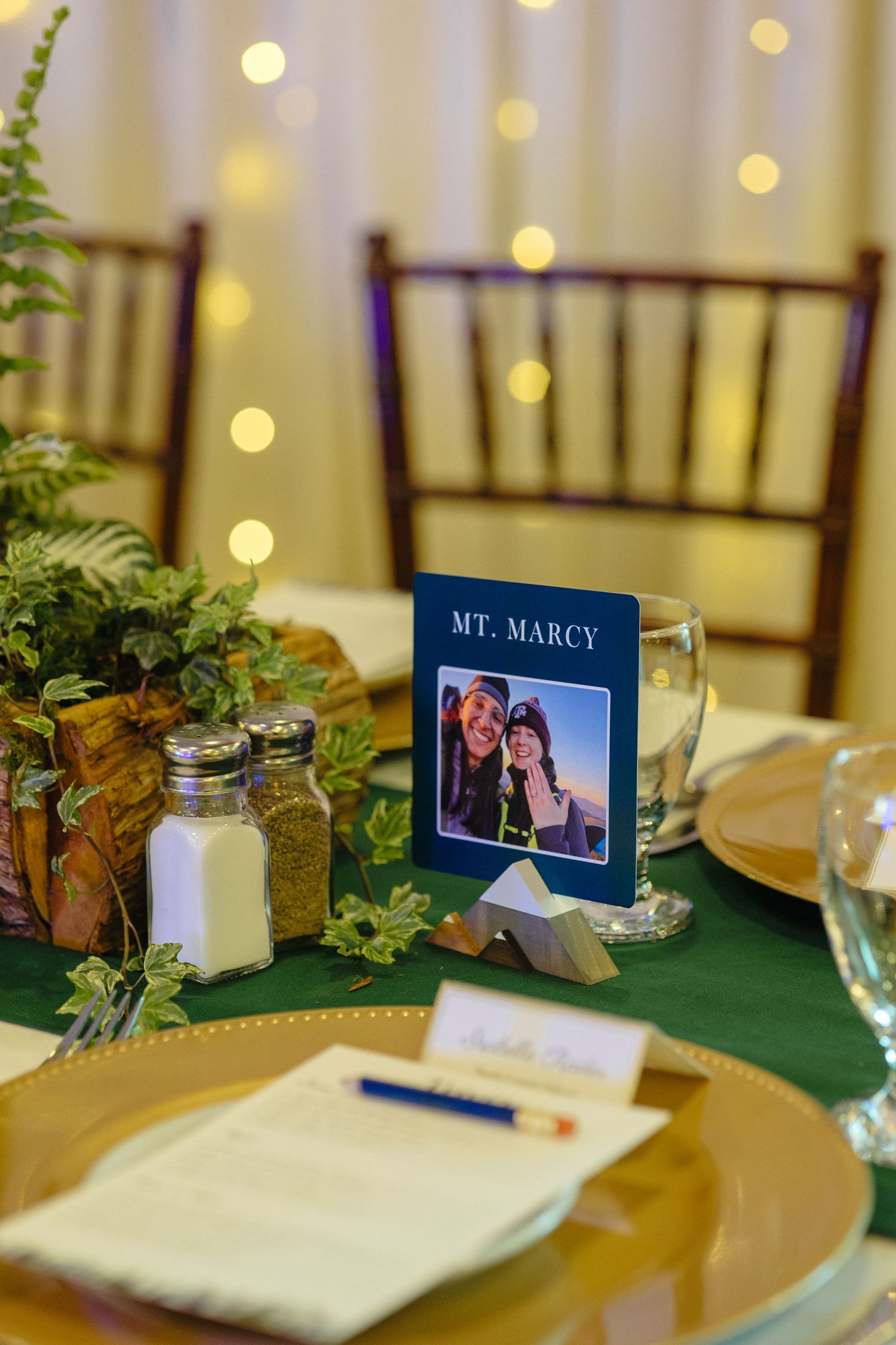 Wedding reception table with Mt. Marcy sign, photo card, greenery centerpiece, gold charger plates, and warm string lights.