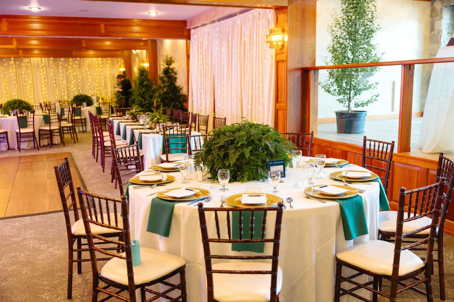Elegant wedding reception venue with round tables, white linens, teal napkins, gold chargers, and lush greenery centerpieces.