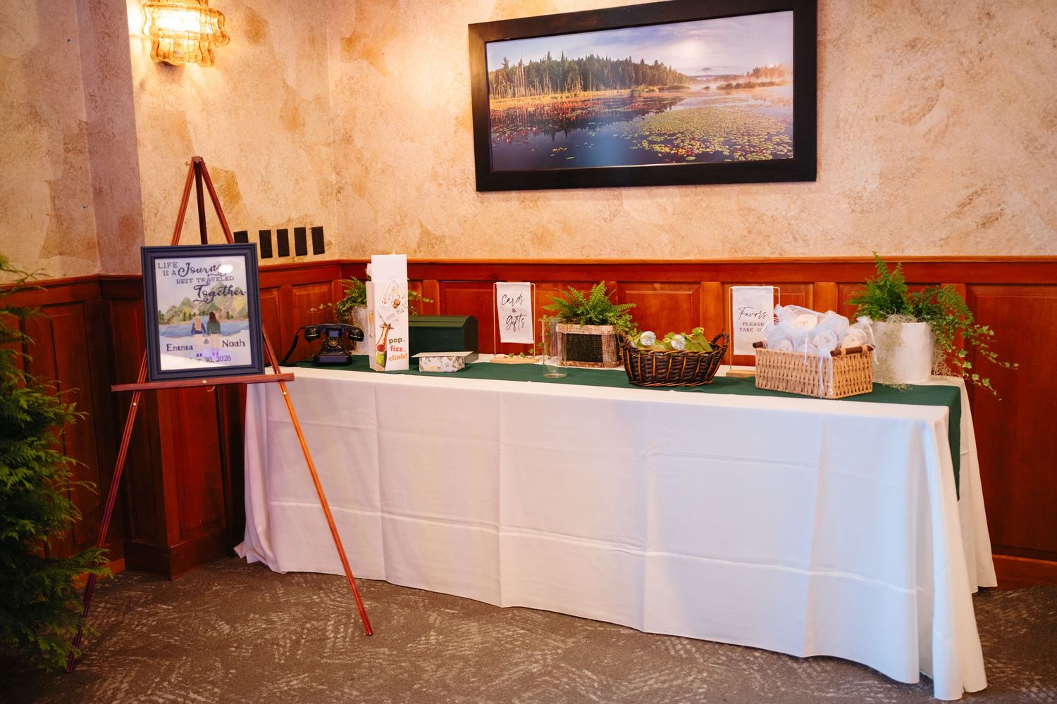 Elegant wedding reception welcome table with white linen, greenery, and landscape painting.