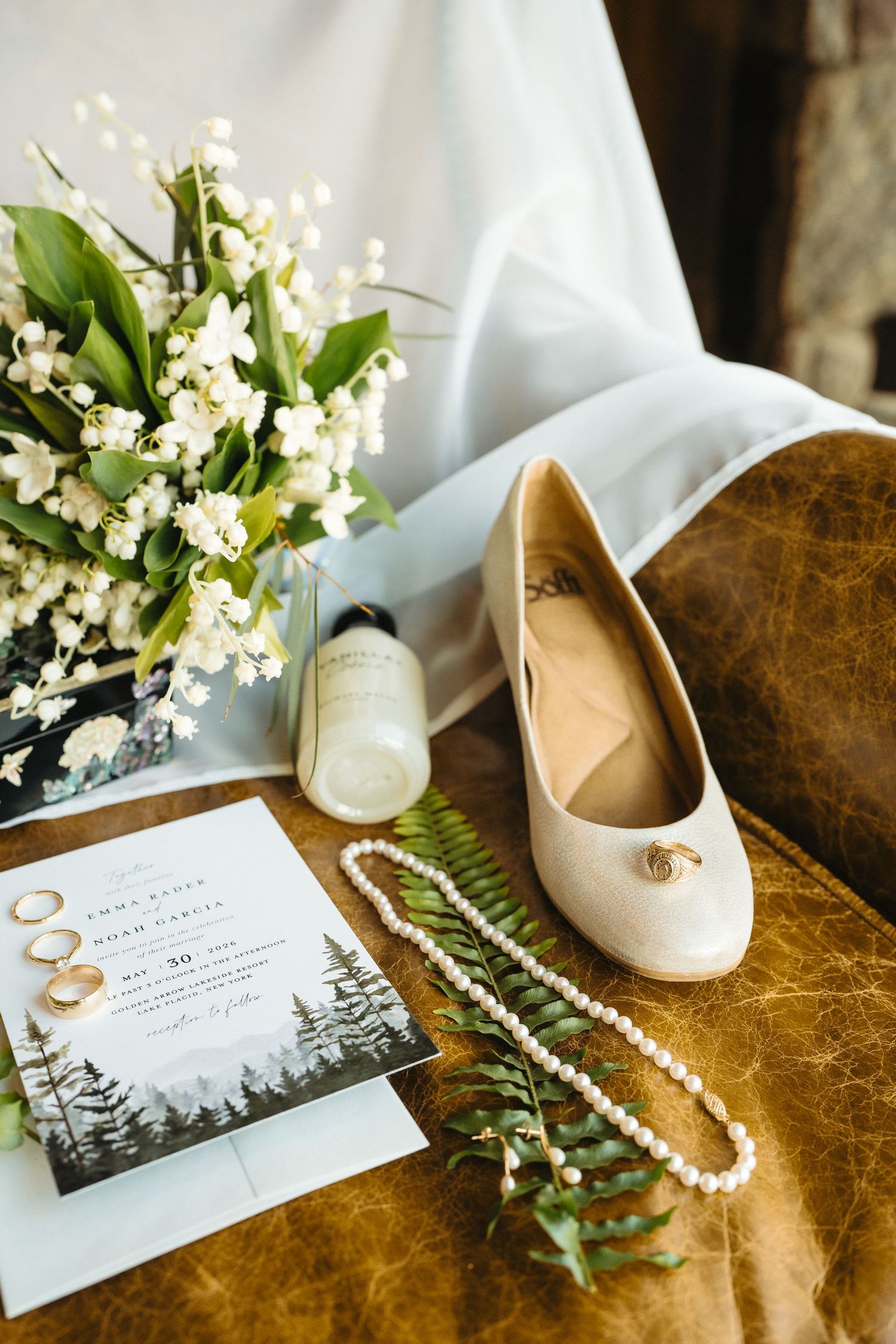 Wedding flat lay with bridal shoes, pearl necklace, rings, invitation, and lily of the valley bouquet on leather surface.