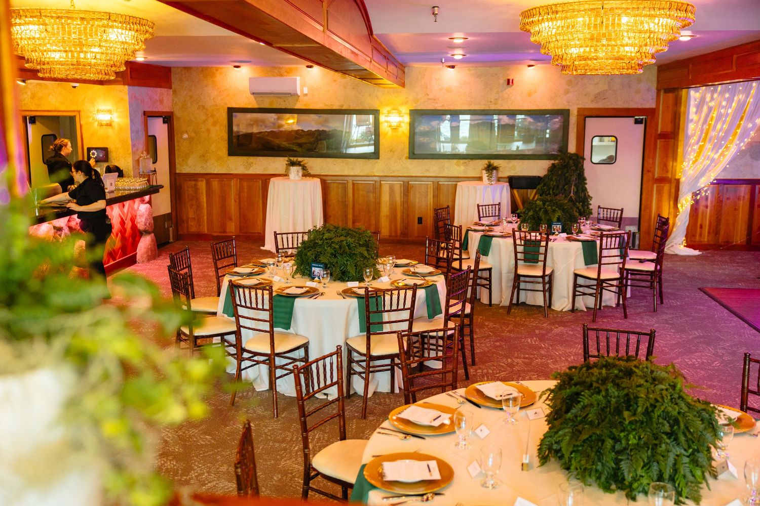 Elegant banquet hall with round tables, chiavari chairs, lush greenery centerpieces, and warm chandelier lighting.